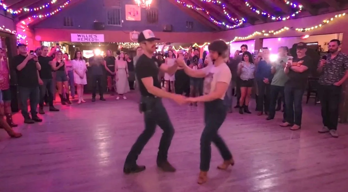 A couple two-stepping together on the dance floor at Sagebrush in Austin, Texas, with a crowd watching under string lights