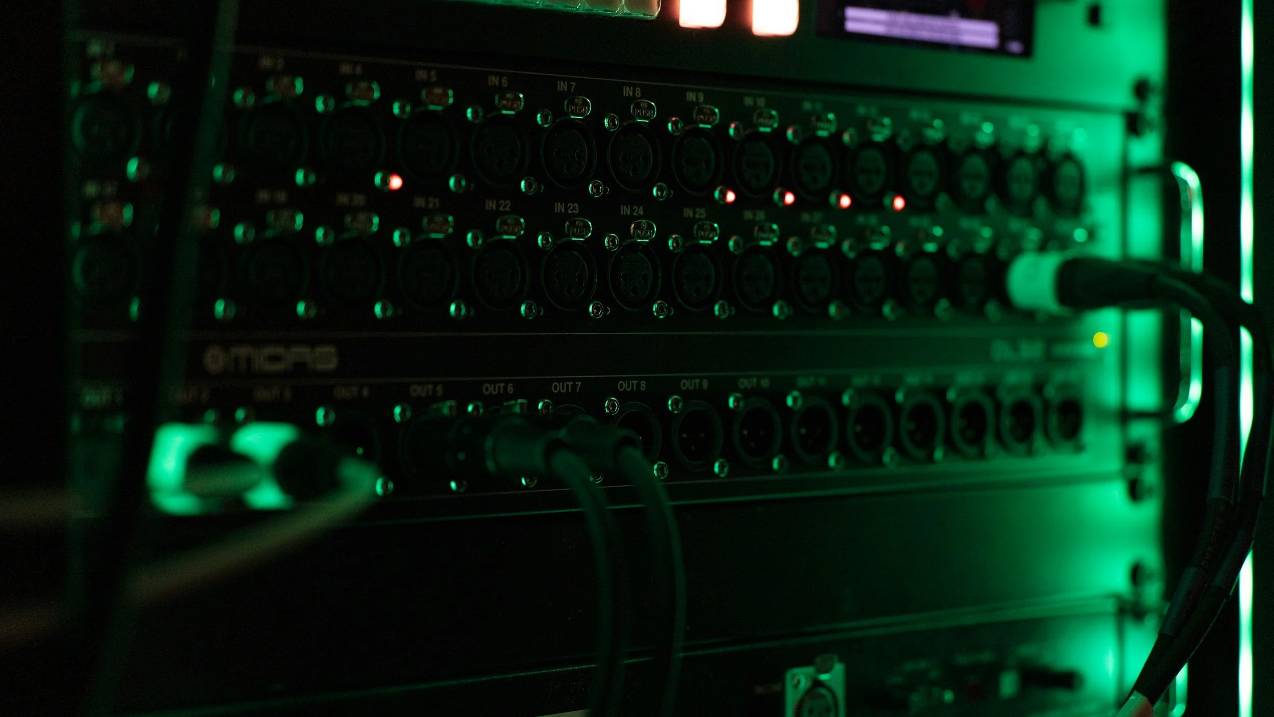 Close-up of a rack with audio or networking equipment, illuminated with green LED lights, with cables connected to the ports.