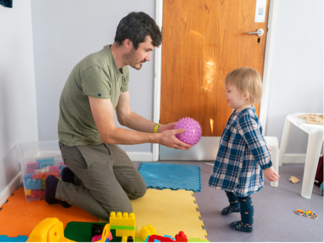 Man kneeling and playing with a young girl in a playroom filled with colorful toys, near a closed wooden door.