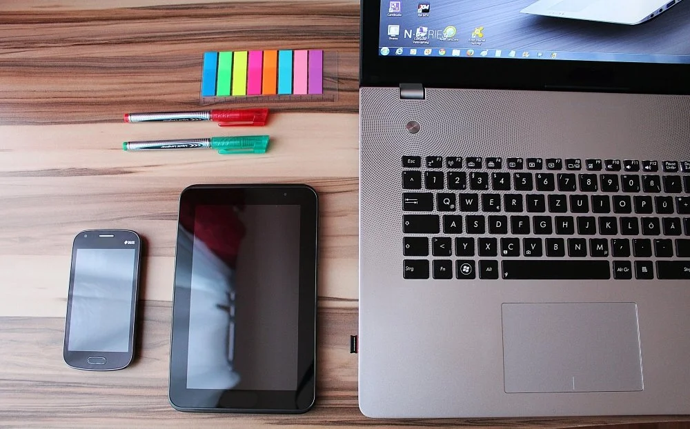 Laptop, smartphone, tablet, colorful sticky notes, and pens on a wooden desk.