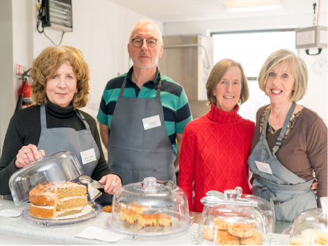 Four people standing behind a table with baked goods under glass covers, in a kitchen or community center. They are smiling and wearing aprons, likely at a bake sale or community event.