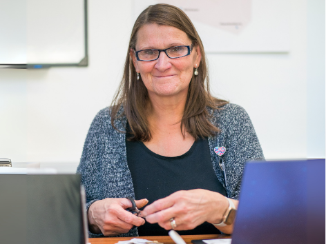 A woman with glasses and shoulder-length brown hair sitting at a desk with a laptop, smiling at the camera.
