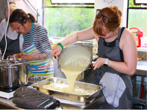 Two women cooking in a kitchen, one is pouring batter into a baking dish while the other is stirring another pan on the stove.