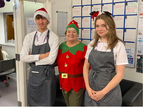 Three people dressed in holiday costumes stand in a room with a calendar and posters on the wall. The person in the middle is dressed as an elf, wearing a red and green outfit with a Christmas hat. The two people on either side are wearing gray aprons, white shirts, and Santa hats.
