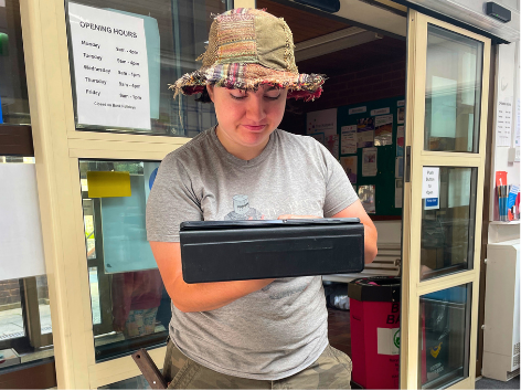 Young woman wearing a hat and gray t-shirt looking at a black device, standing near a door with open windows and a sign with opening hours in the background.