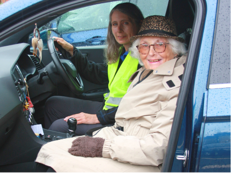 An elderly woman and a young woman sitting in a parked car. The elderly woman is in the driver's seat, wearing a beige coat, leopard print hat, and glasses. The young woman is in the passenger seat, wearing a neon yellow safety vest and smiling.
