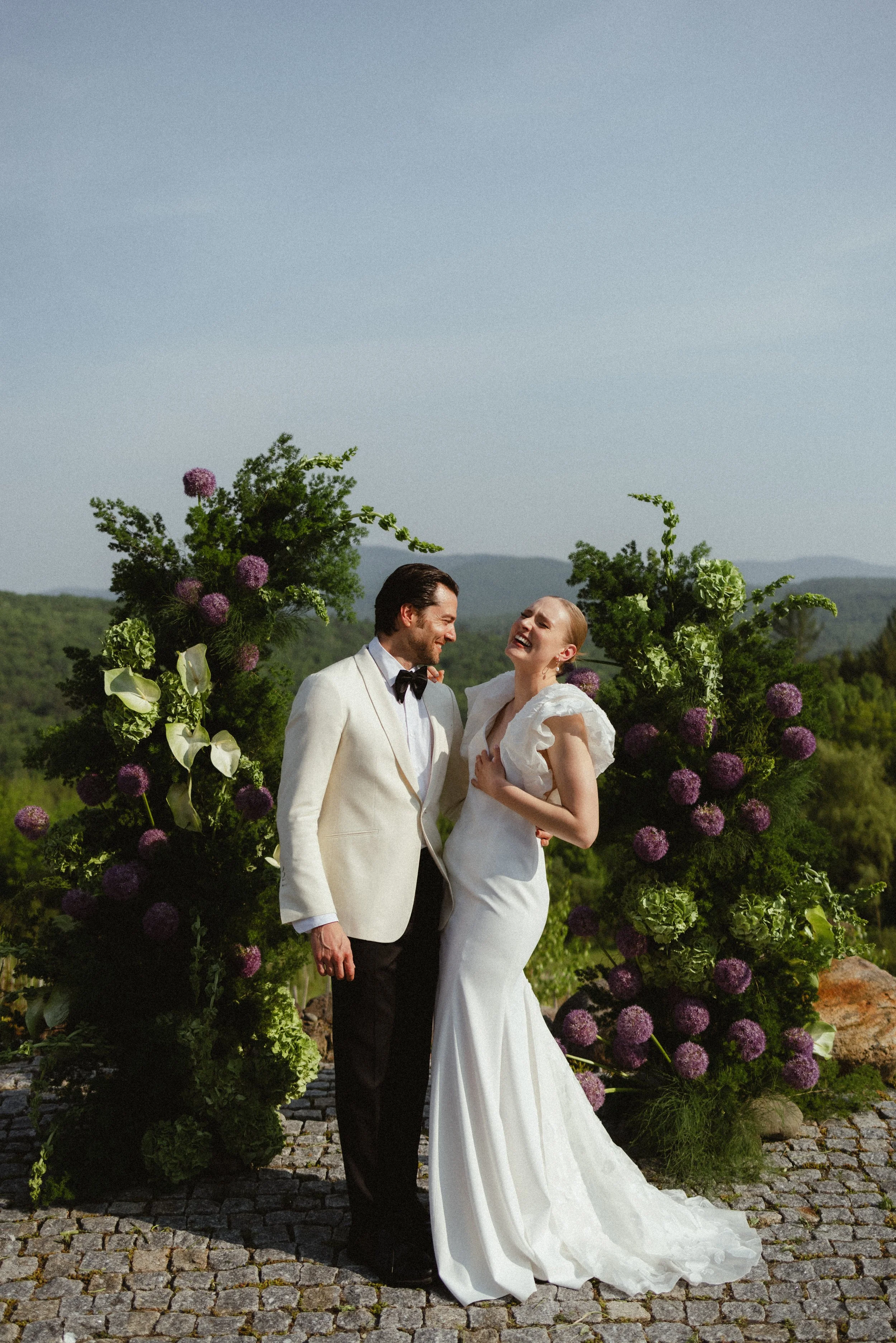 A laughing bride and groom standing in front of an outdoor floral arrangement.