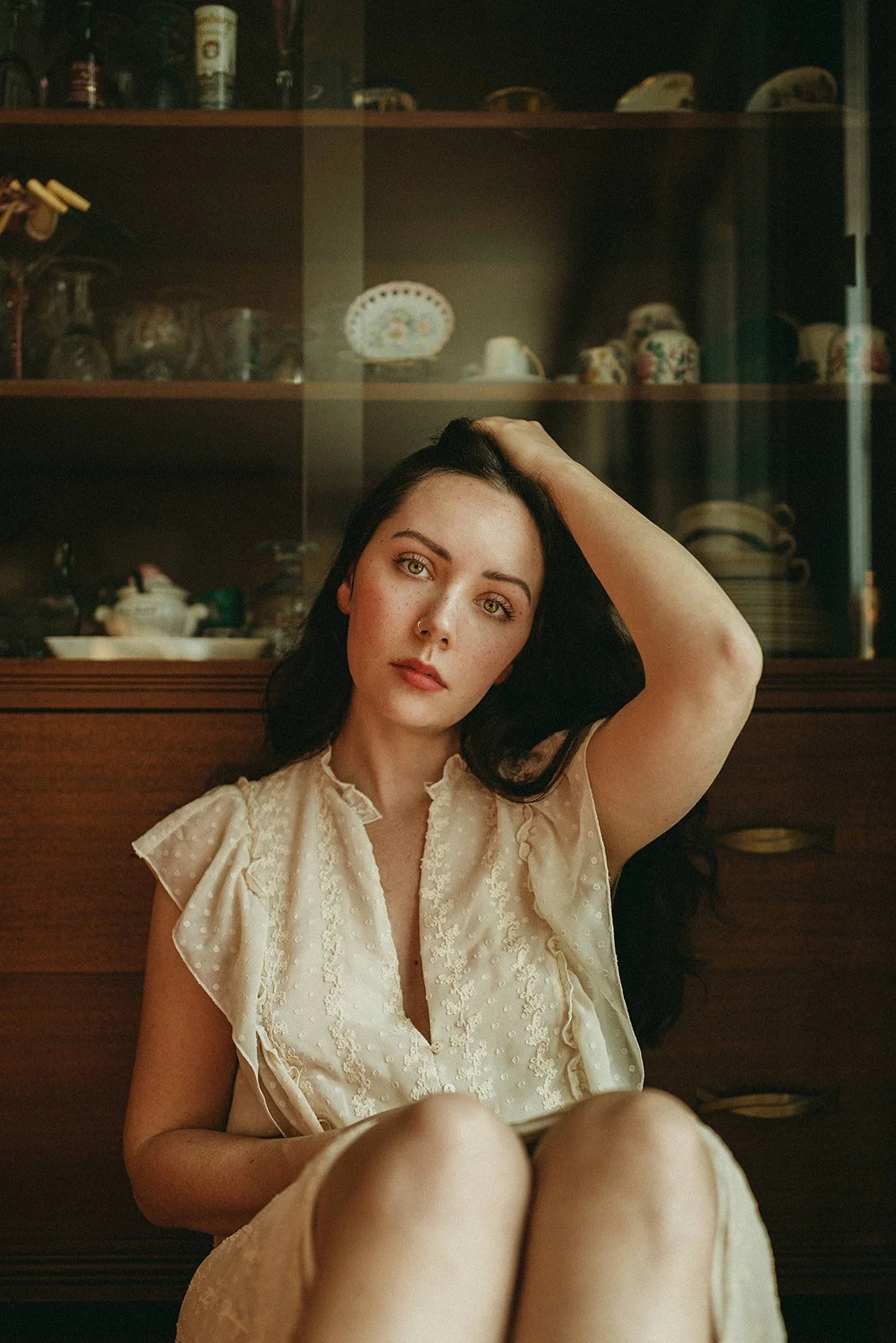 A young woman with long black hair and green eyes sitting in front of a wooden cabinet filled with dishes and decorative items, wearing a cream-colored, textured blouse, with one hand on her head and looking directly at the camera.