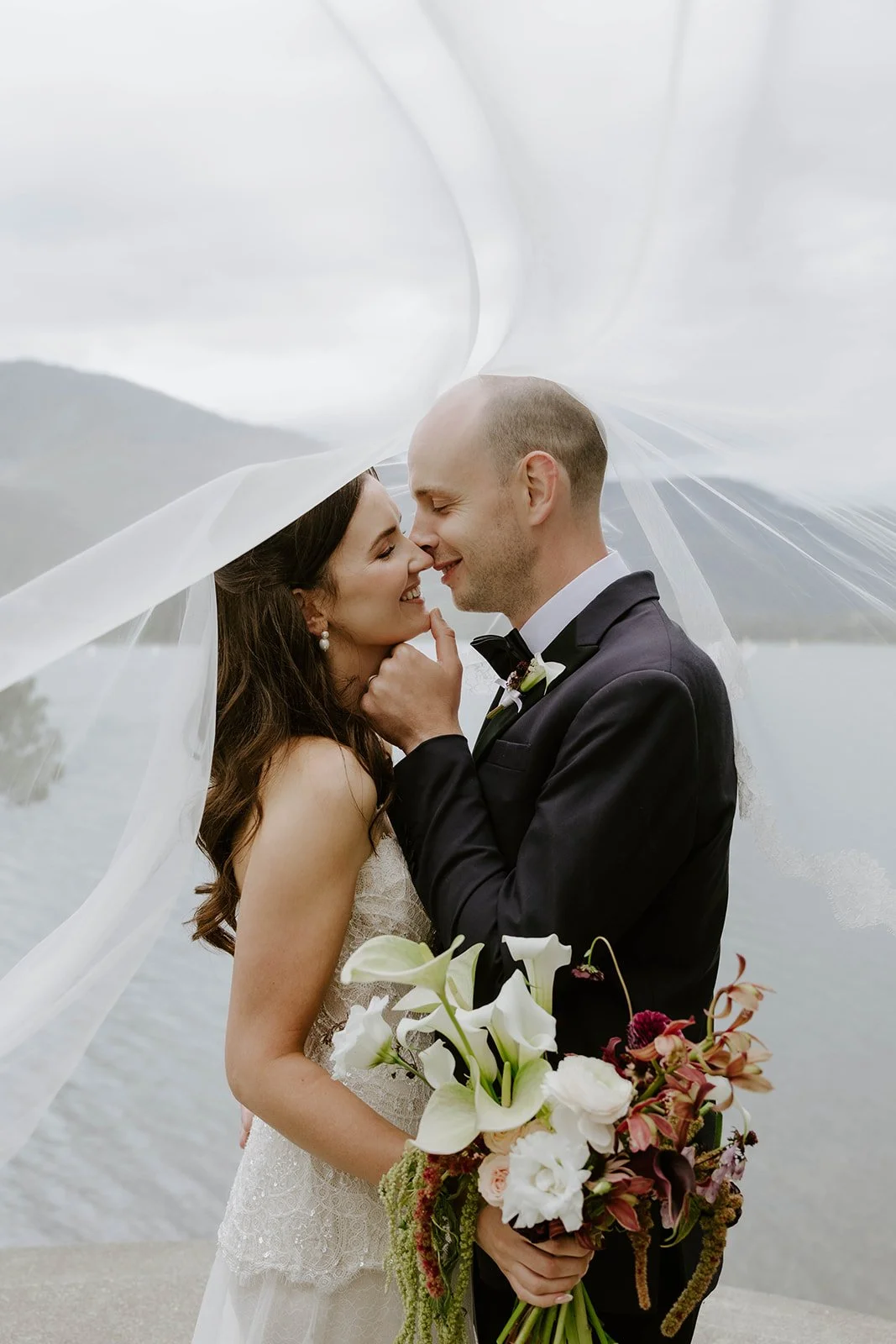 A newlywed couple shares an intimate moment under the bride's veil, with the bride holding a bouquet of white calla lilies and other flowers. They are outdoors near a body of water with mountains in the background, overcast sky.