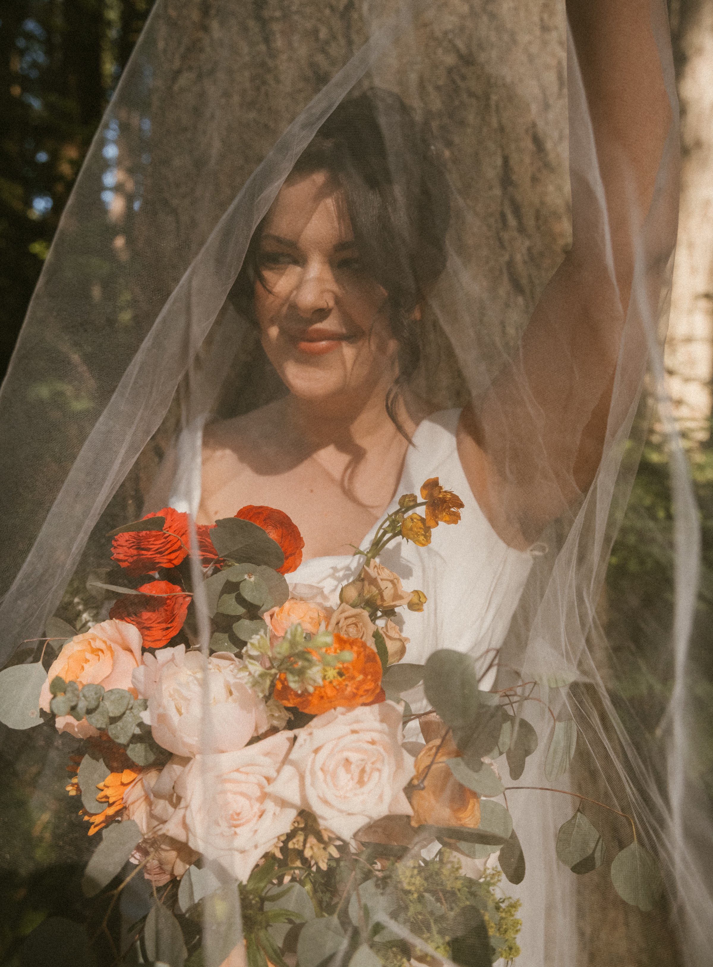 A woman with dark hair and a nose piercing behind a sheer veil, holding a bouquet of peach, cream, and orange flowers with eucalyptus leaves, outdoors near a tree.