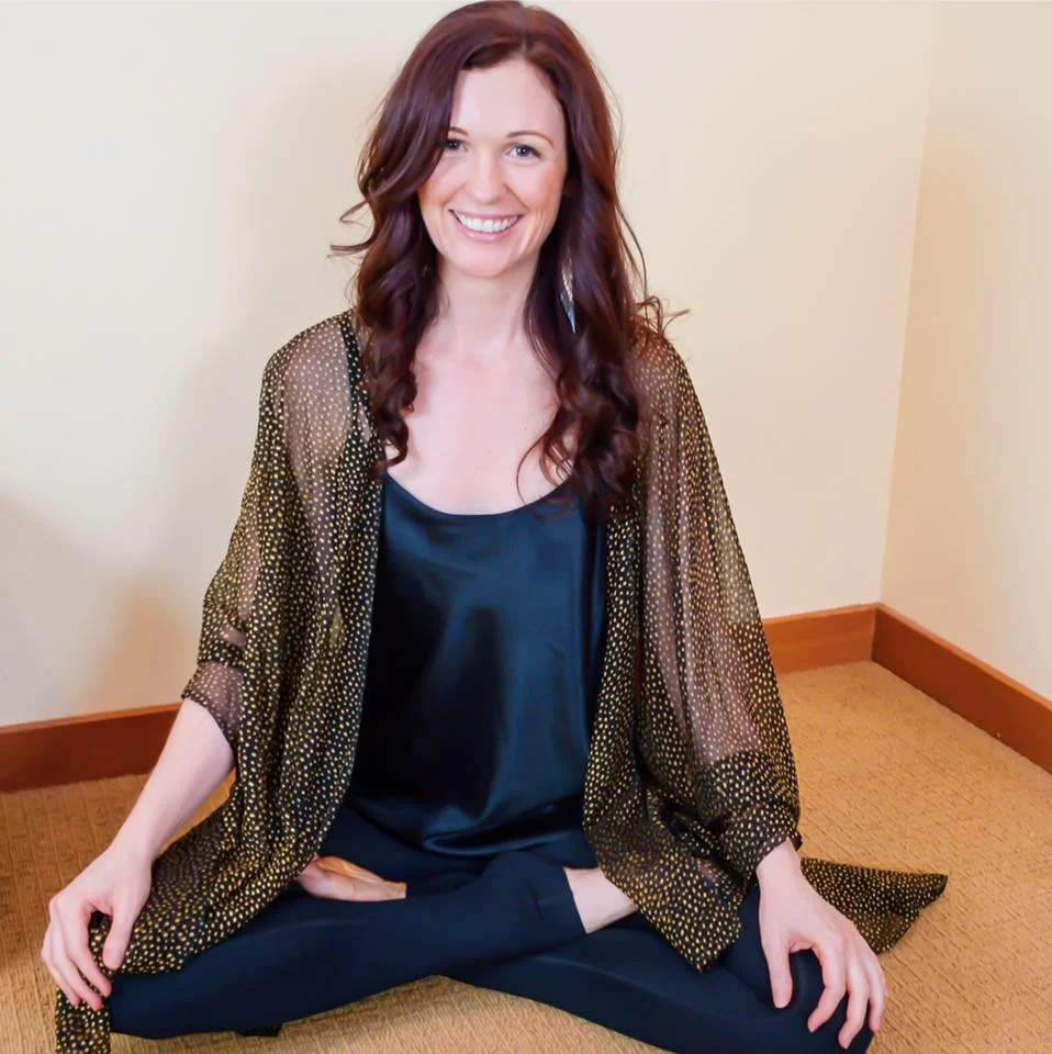 Person sitting cross-legged on carpet, smiling, wearing a sheer black and gold outfit in a bright room.