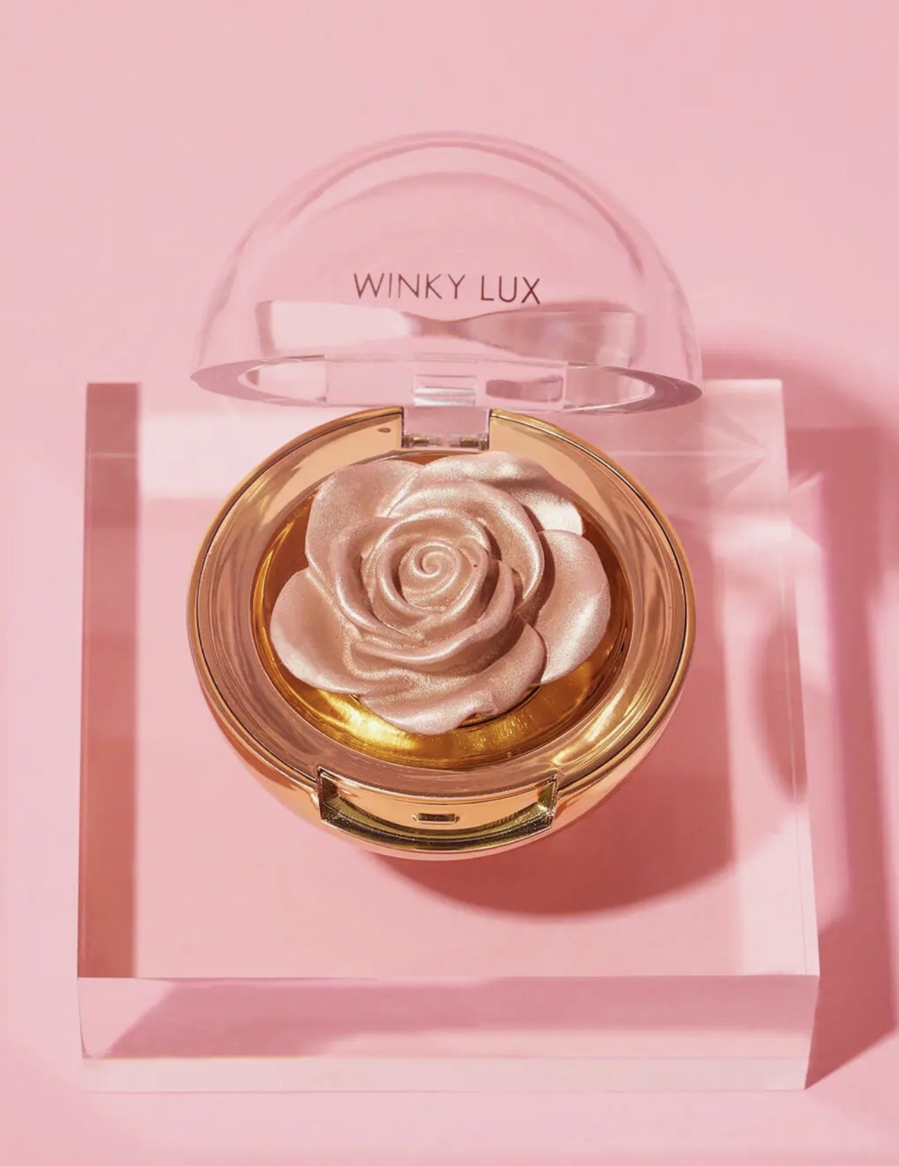 A pink compact with a rose-shaped design inside, placed on a matching pink surface. In the background, there is a clear plastic lid with the text "WINKY LUX".