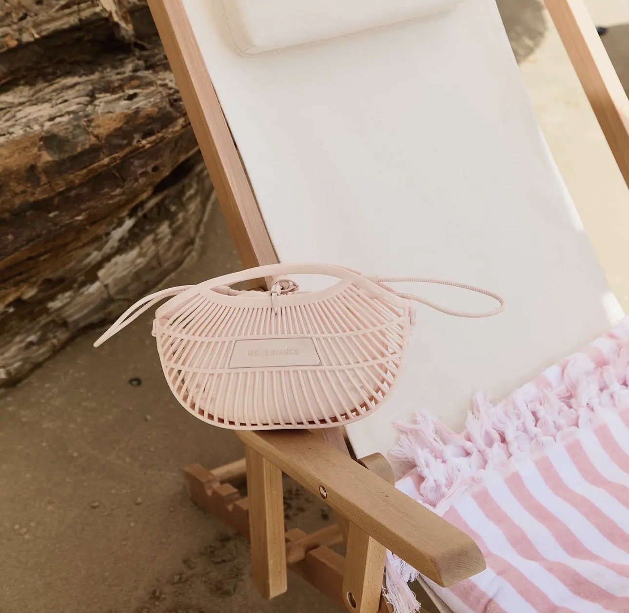 A pink, modern, slatted jelly Melie Bianco handbag resting on a wooden beach chair with a pink and white striped towel, next to a large stone wall.
