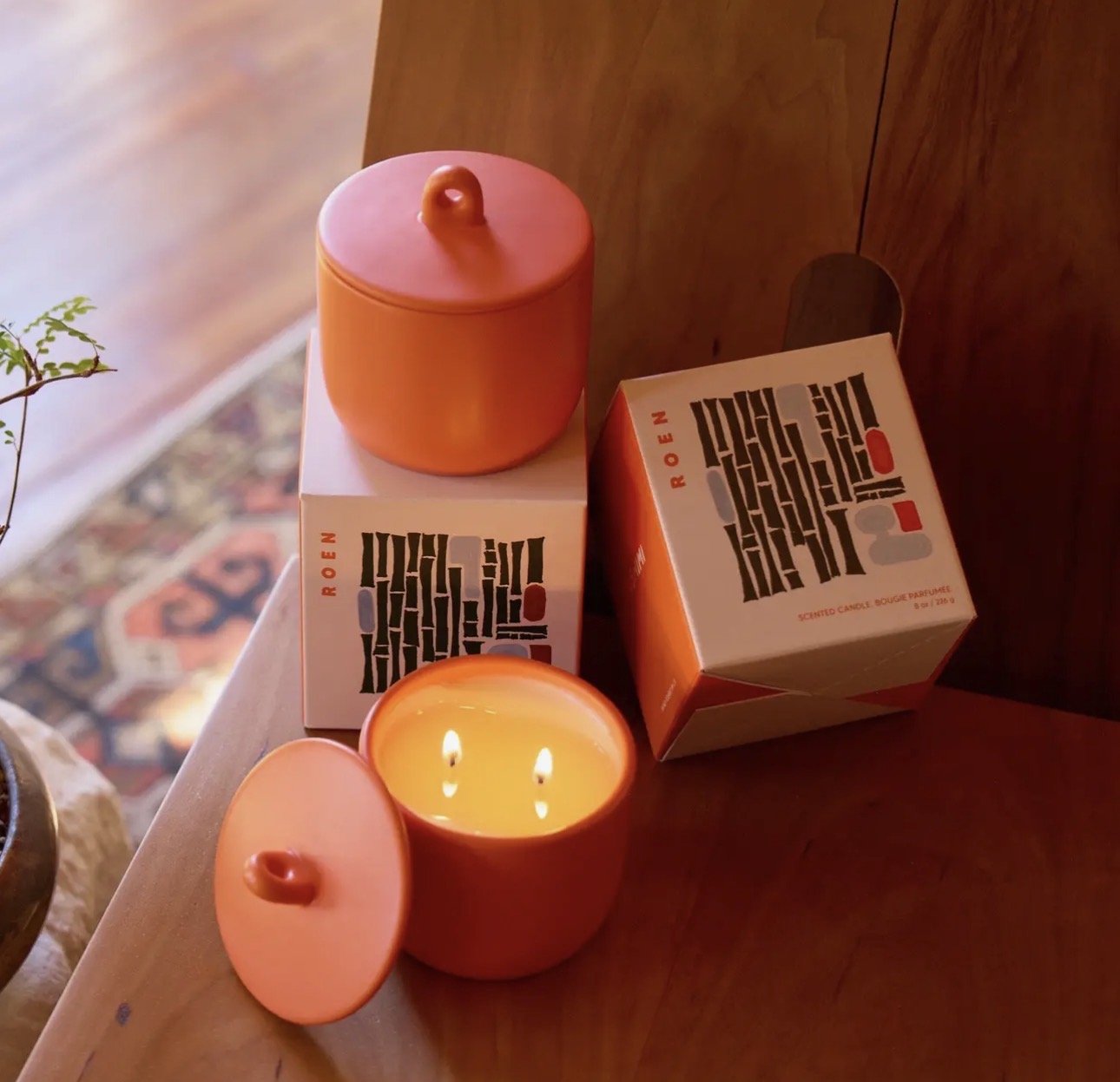 A lit scented candle in an orange container with its lid beside it. Two boxes of scented candles labeled 'ROEN Fushimi' are on a wooden surface, one upright and one tilted. A small plant and a patterned rug are partially visible in the background.