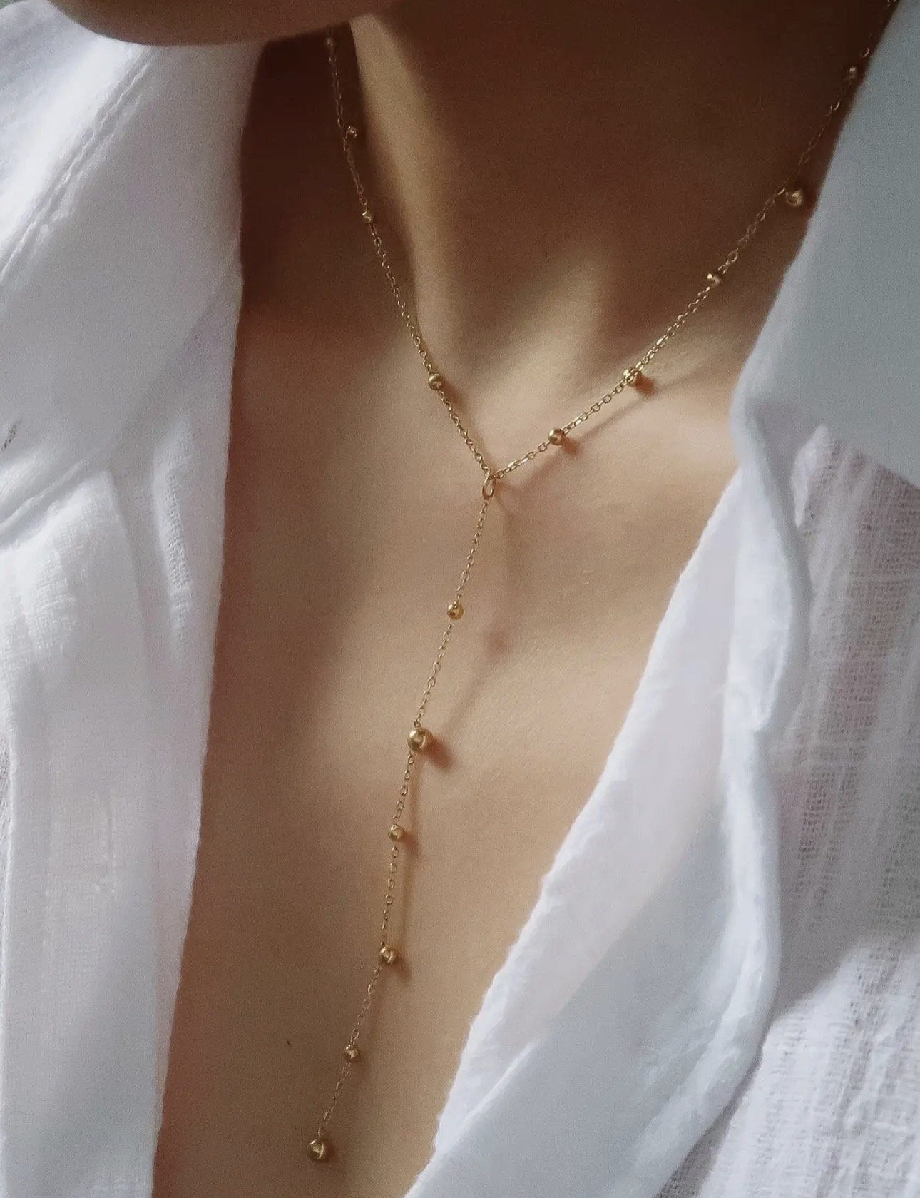 Close-up of a woman wearing a gold layered necklace with small beads, partially visible white shirt, and skin showing at the neck and chest.