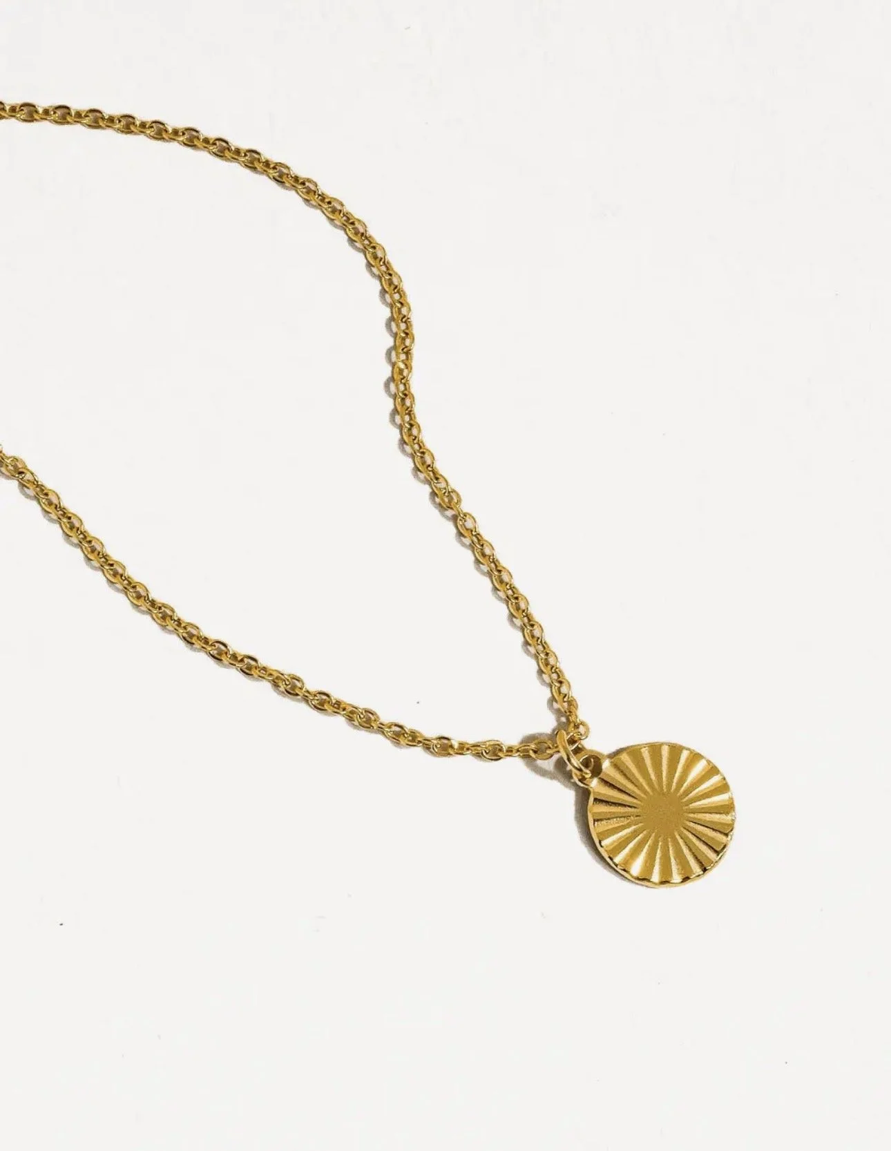Gold necklace with a circular pendant featuring a sunburst design on a white background.