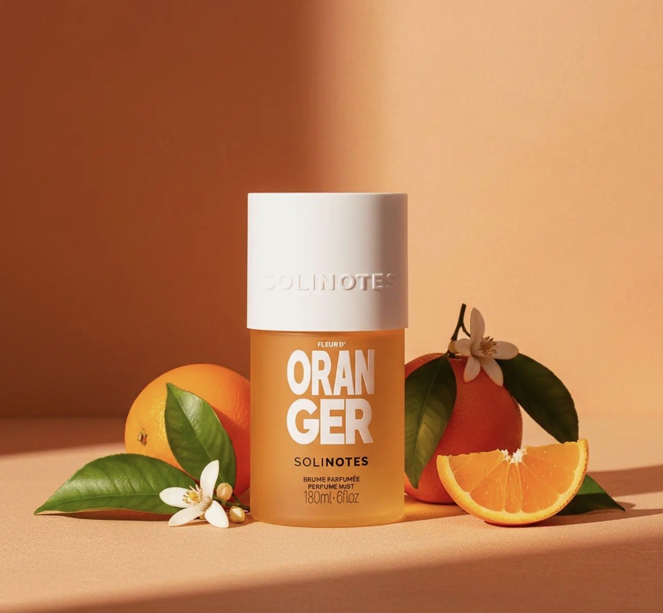 A perfume mist bottle labeled 'Solnotes Oranger' surrounded by oranges, orange slices, green leaves, and orange blossoms against a peach-toned background.