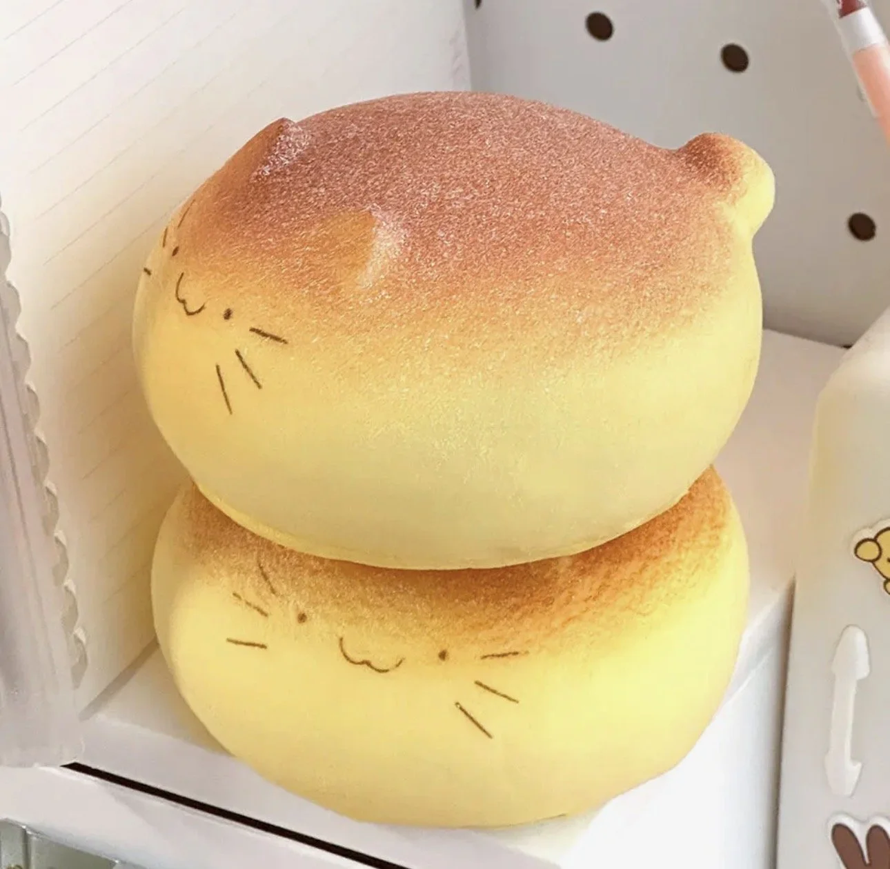 Two round plush toys designed to look like sleeping cats, yellow with closed eyes, rosy cheeks, and small ears, placed in a display case.