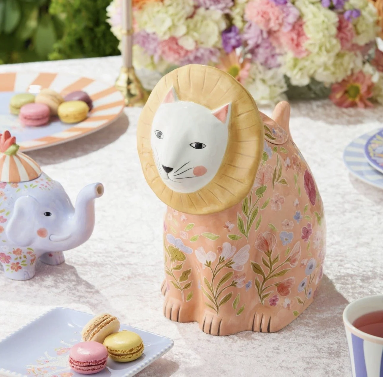 Decorative ceramic animal figurines, including a lion with a human face and a painted elephant, on a table with pastel-colored macarons and floral arrangements in the background.