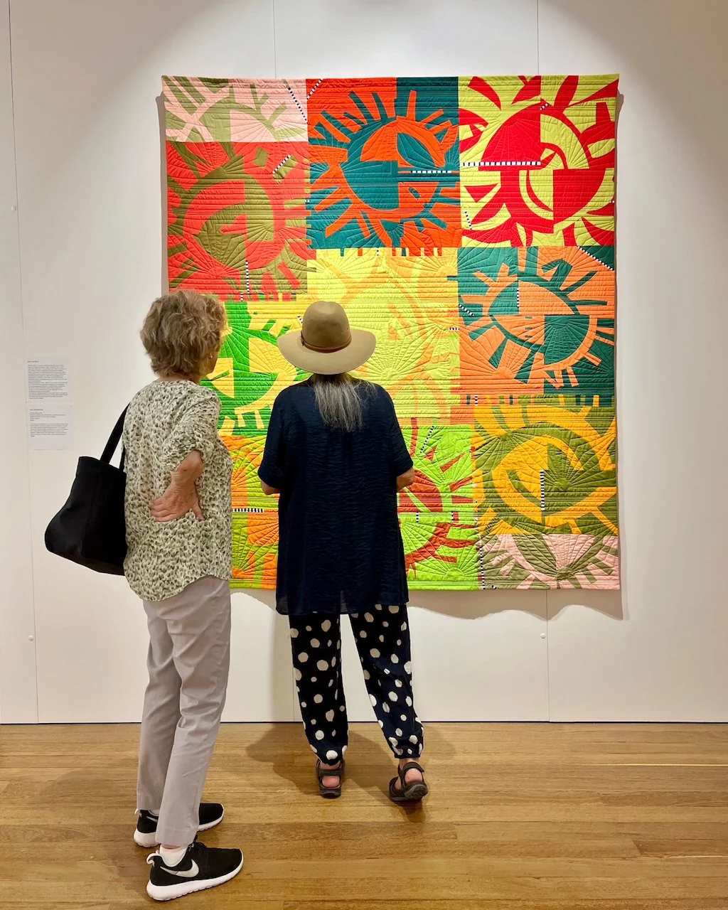 Two women standing looking at a colourful improv quilt hanging on a wall.