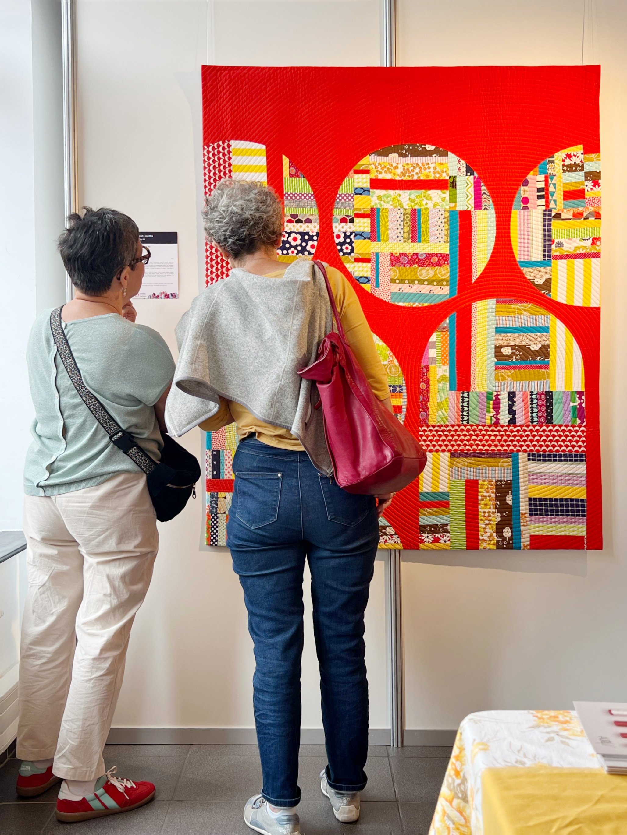 Two women looking at a red quilt with large circular shapes cut out of it to reveal colourful scrappy piecing