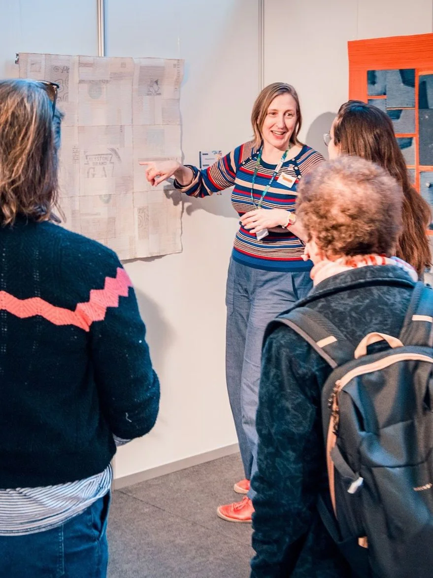 Two women facing the artist and her interpreter talking about the two quilts hanging on the wall