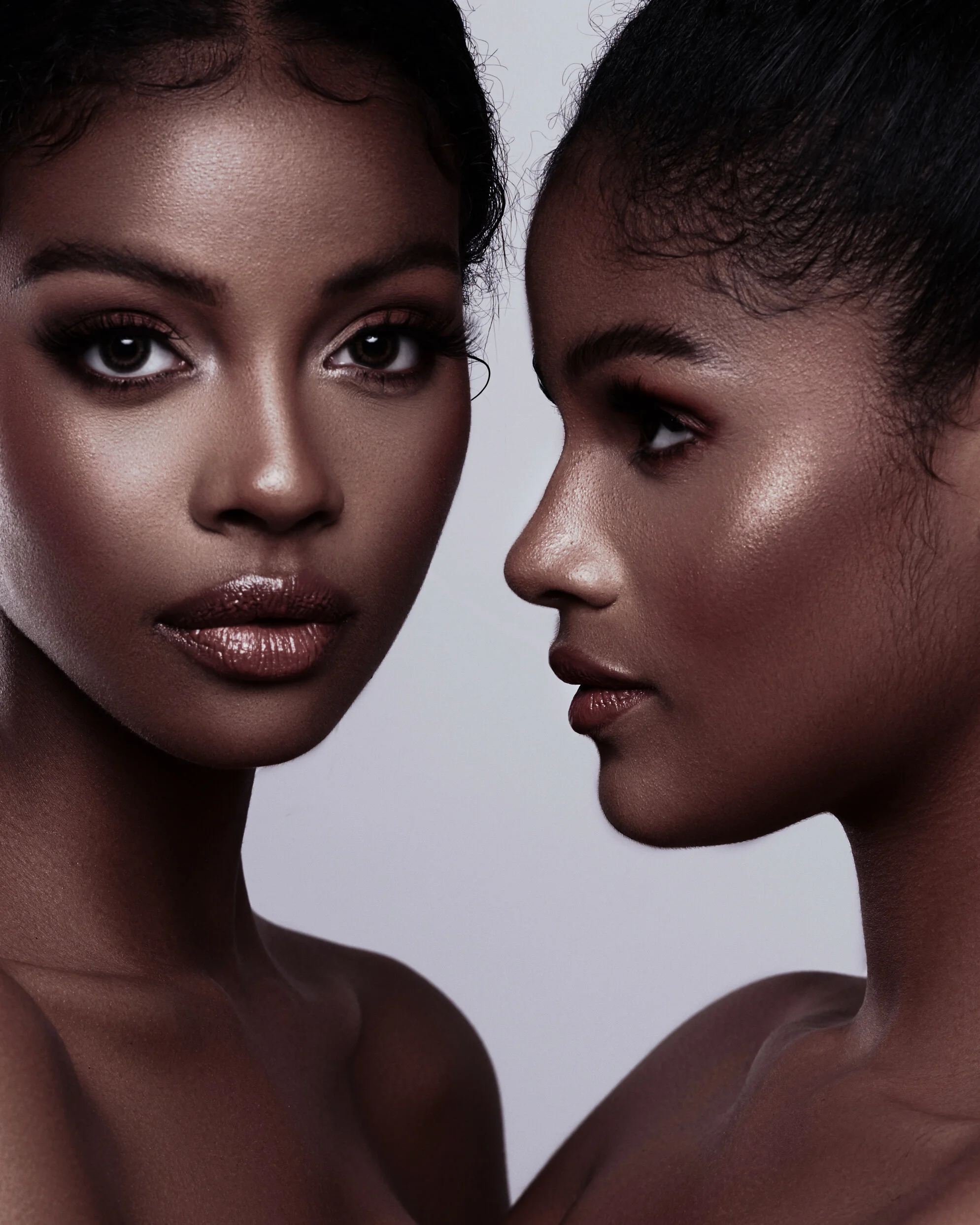 Close-up portrait of two women with dark skin tones, facing each other, with one woman looking directly at the camera and the other in profile.