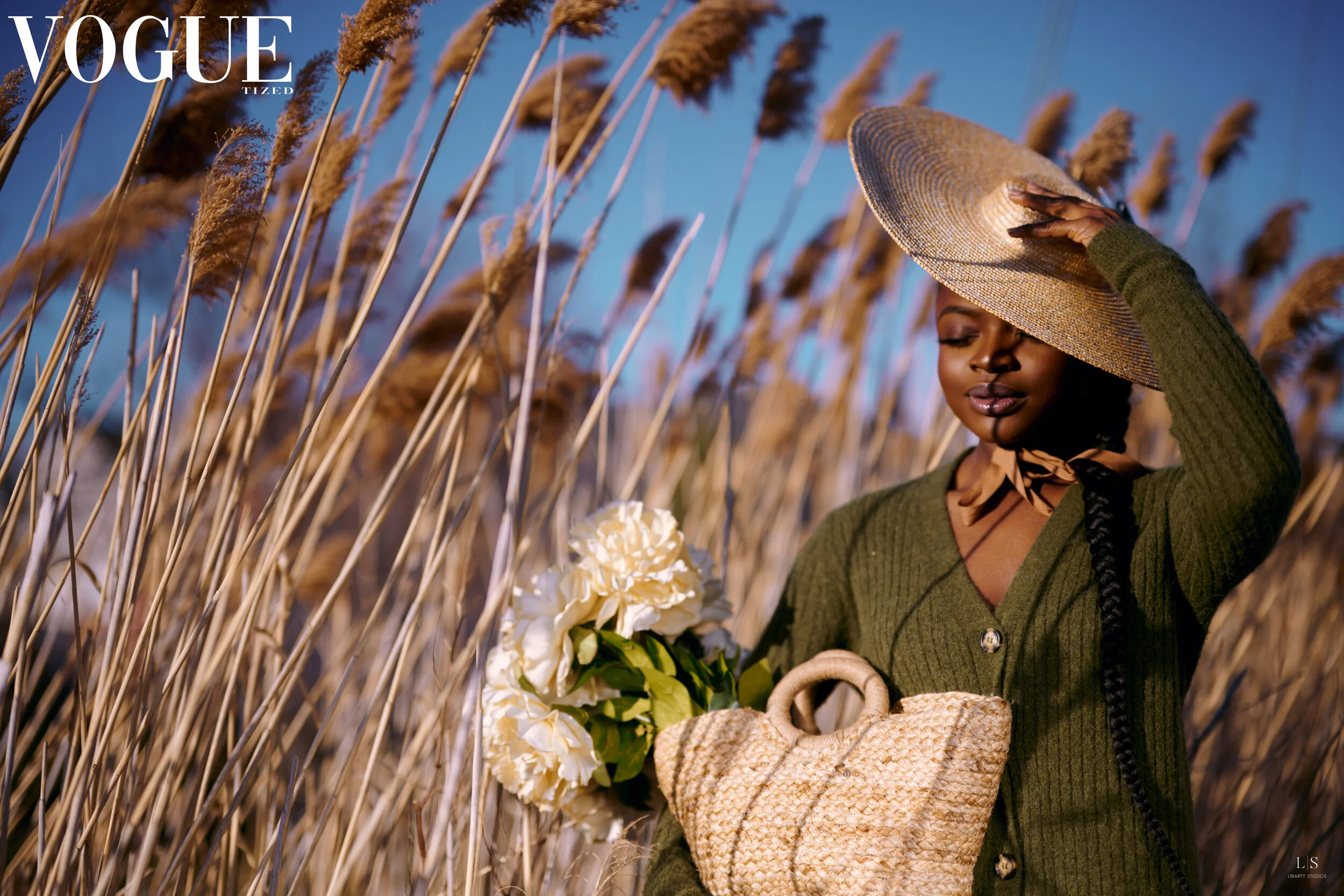 A woman in a green cardigan holding a bouquet of white flowers, standing in a field of tall brown reeds, wearing a large straw hat, with her eyes closed and hand on the hat, during what appears to be late afternoon or early evening.