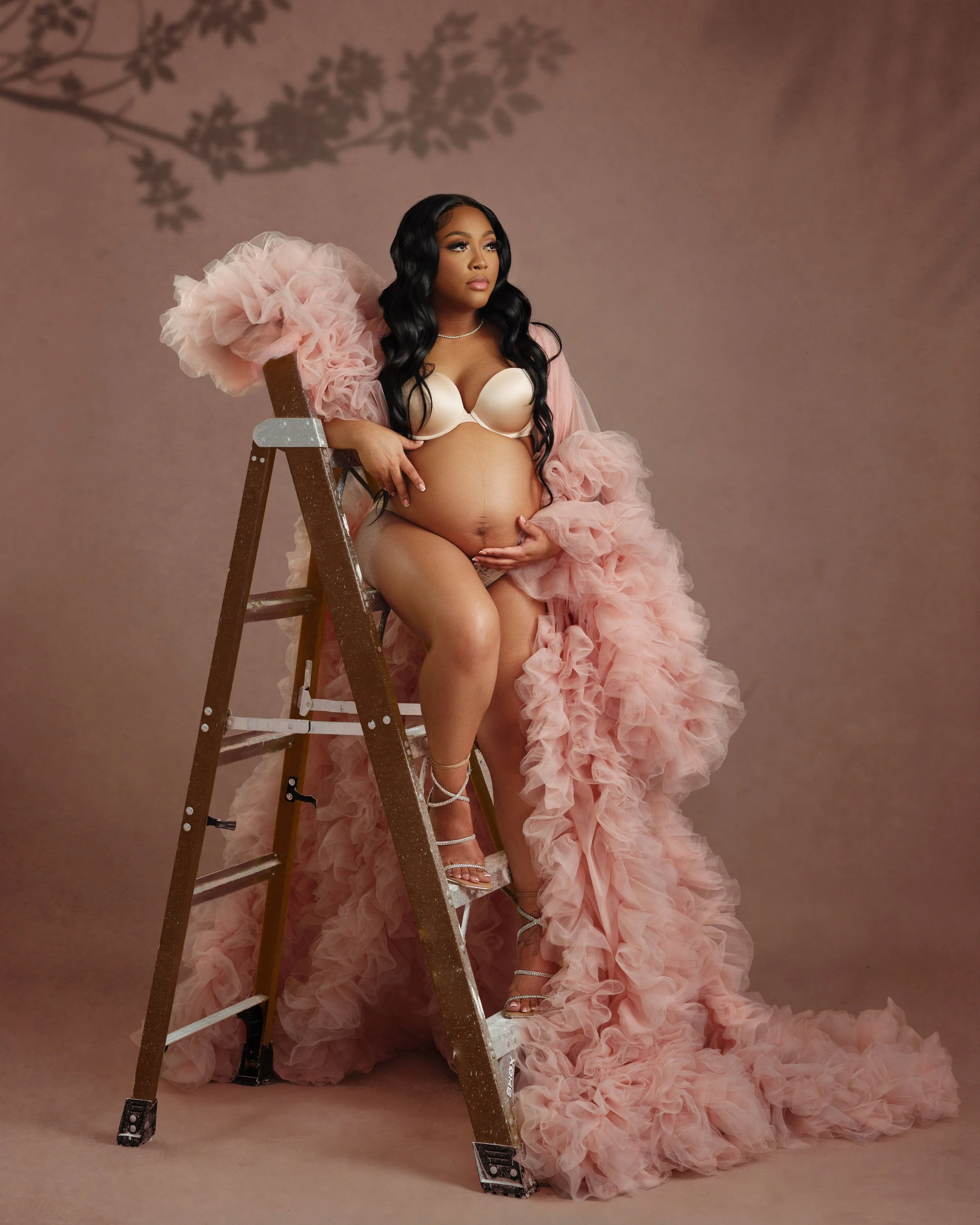 A pregnant woman with long black hair, wearing a cream bra, silver high heels, and a pink ruffled gown, is sitting on a ladder with one foot on the step and the other on the ground. She is gently holding her belly with one hand and resting her other 