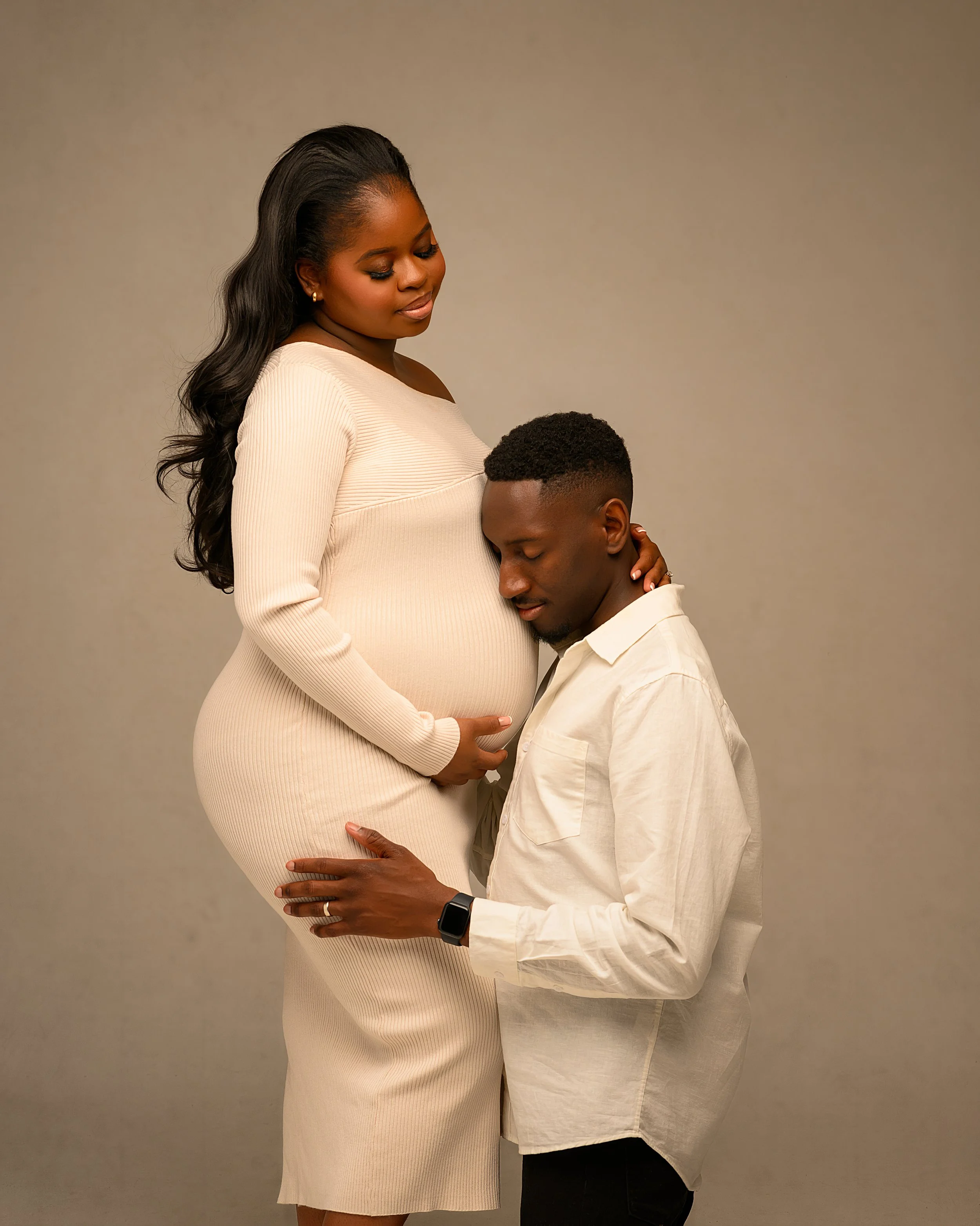A pregnant woman in a cream dress standing next to a man in a white shirt with her hand on her belly, and the man kneeling with his head to her belly, both with their eyes closed, in a neutral setting.