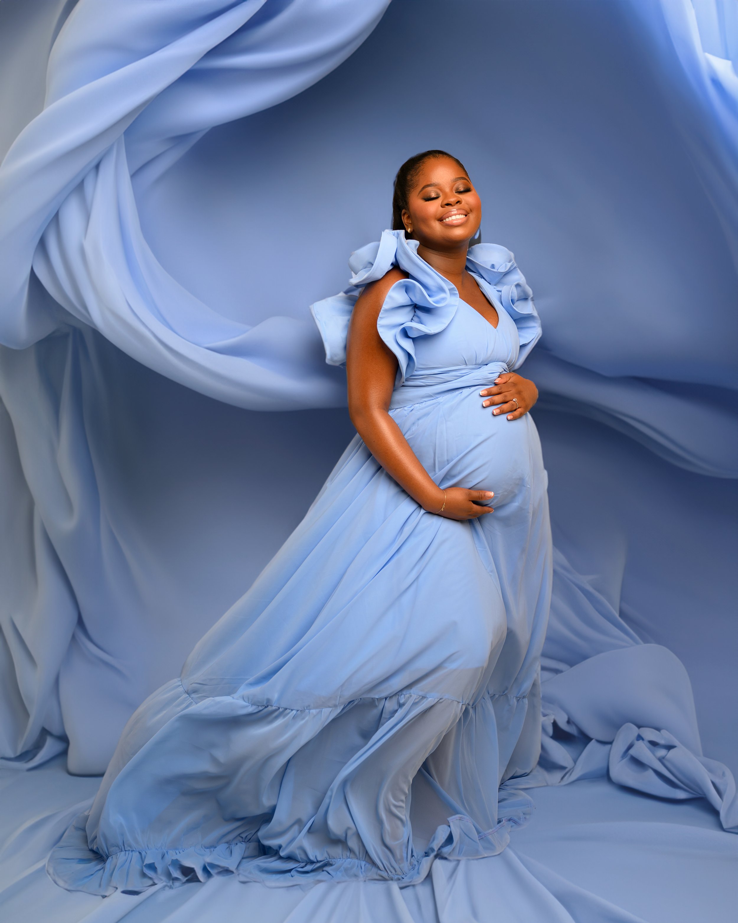 A pregnant woman in a long blue dress with ruffled sleeves, standing in front of a blue fabric backdrop, smiling with her eyes closed and hand on her belly.