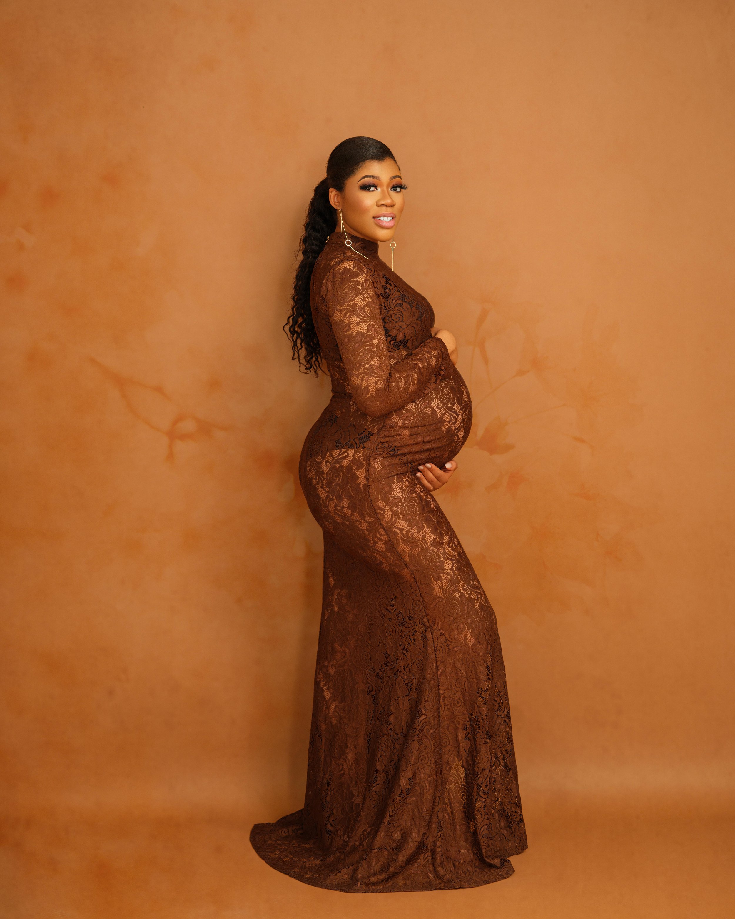 A pregnant woman in a long, brown lace dress standing against a matching brown background, holding her belly and smiling at the camera.