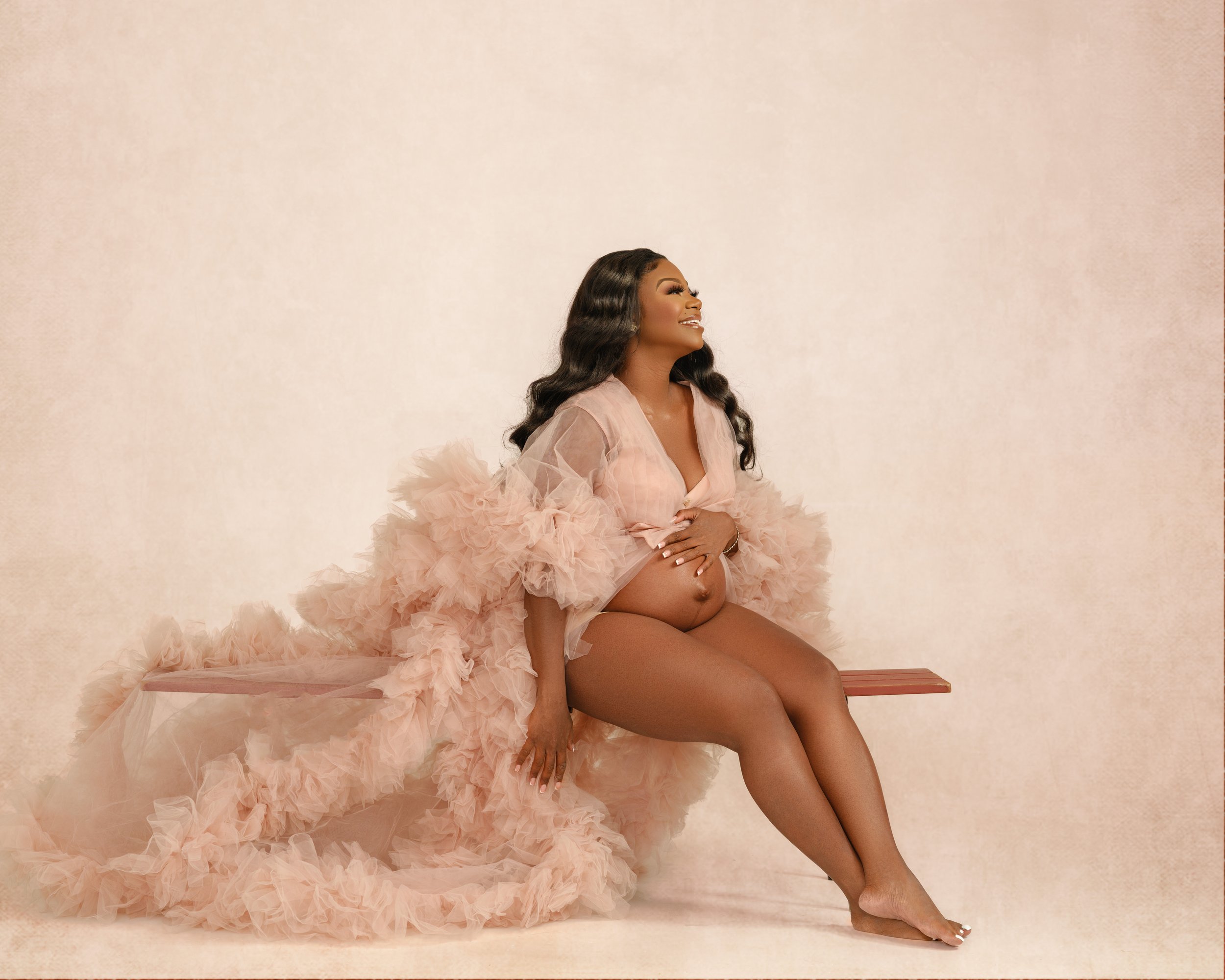 Pregnant woman sitting on a wooden bench, wearing a flowing pink ruffled gown, smiling with her eyes closed, against a plain light-colored background.