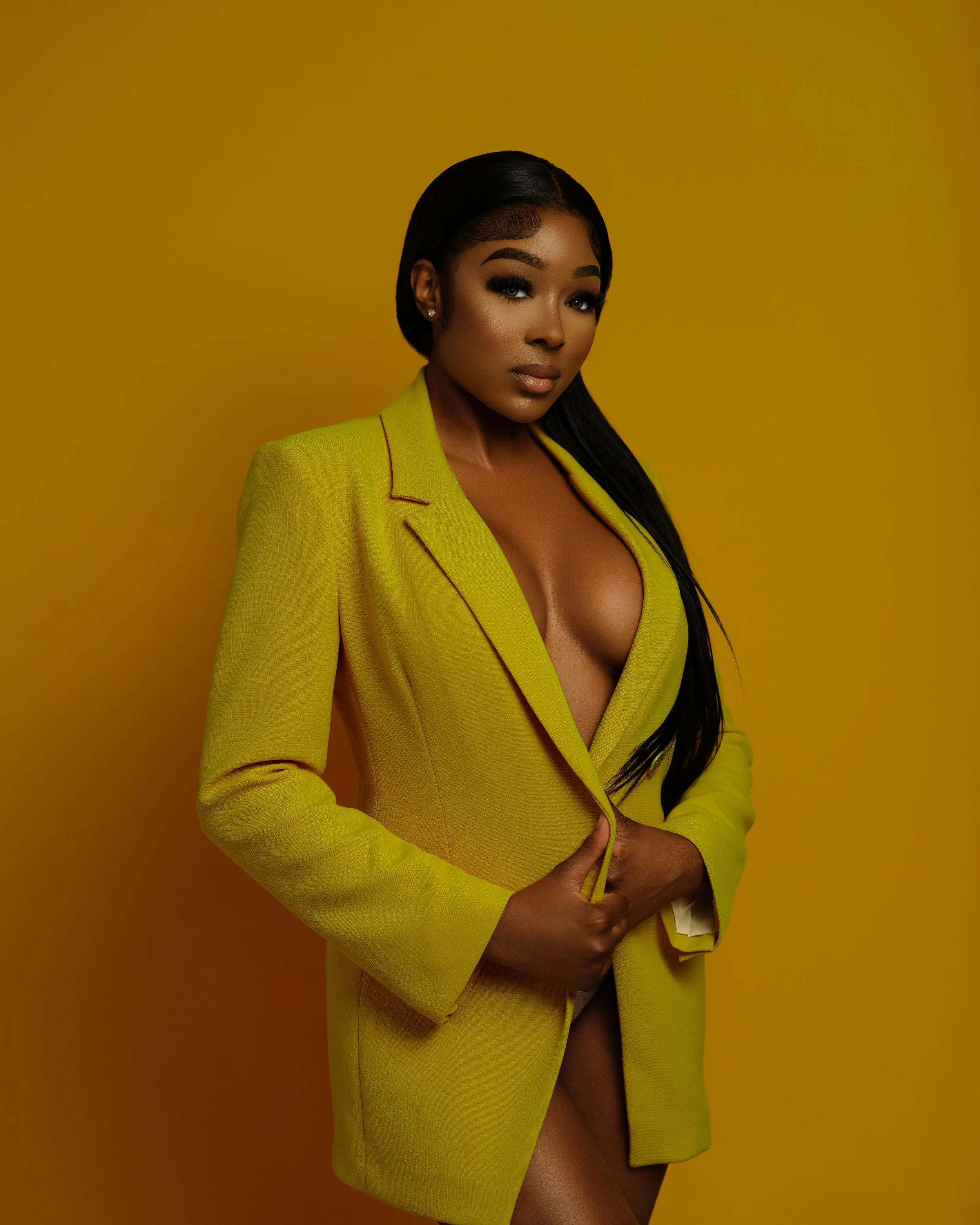A woman with dark hair styled straight, wearing makeup, dressed in a bright yellow blazer with no shirt underneath, standing against a matching yellow background.