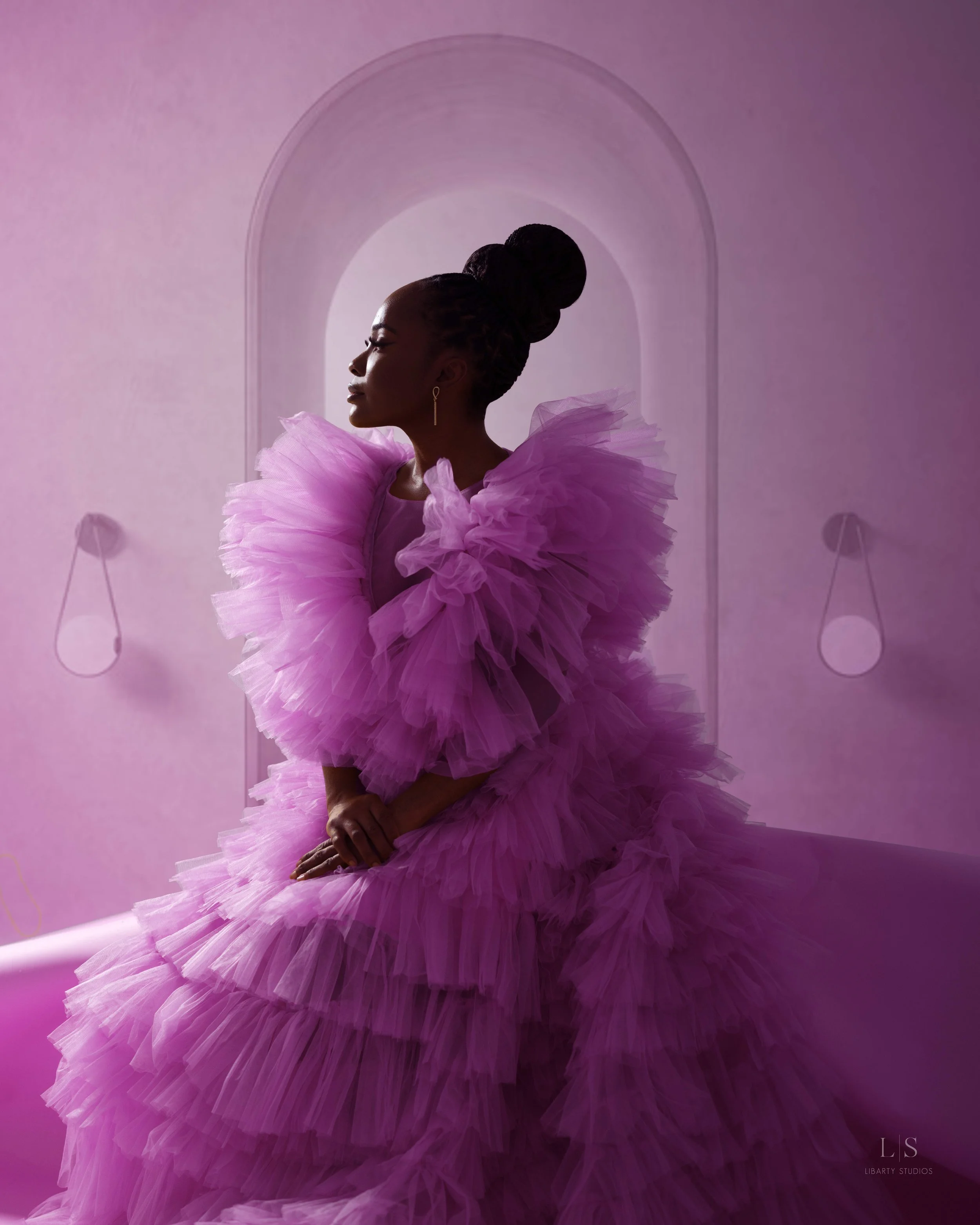 A woman in a voluminous pink ruffled dress sitting with her eyes closed and hands clasped. The background is pink with an arched outline and two wall sconces.