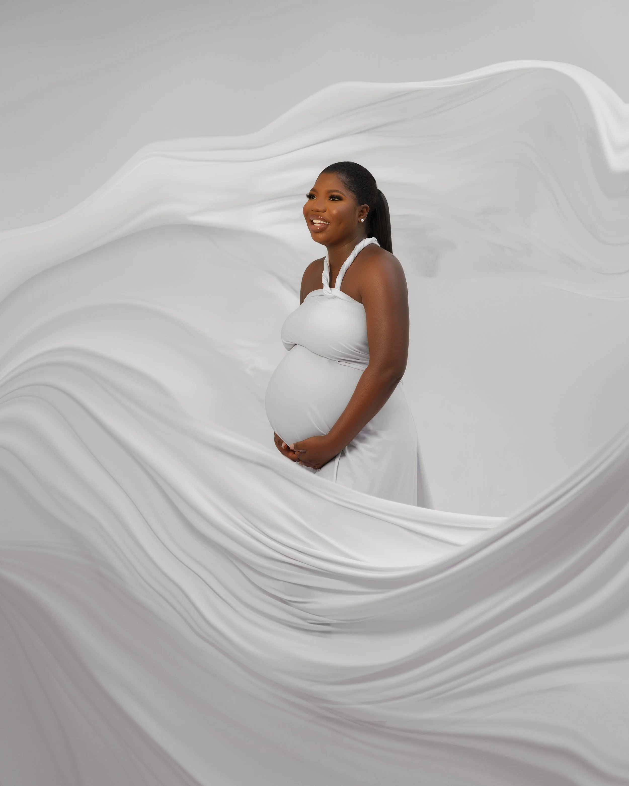 Pregnant woman in a white dress smiling, standing against a white abstract background with flowing fabric-like textures.