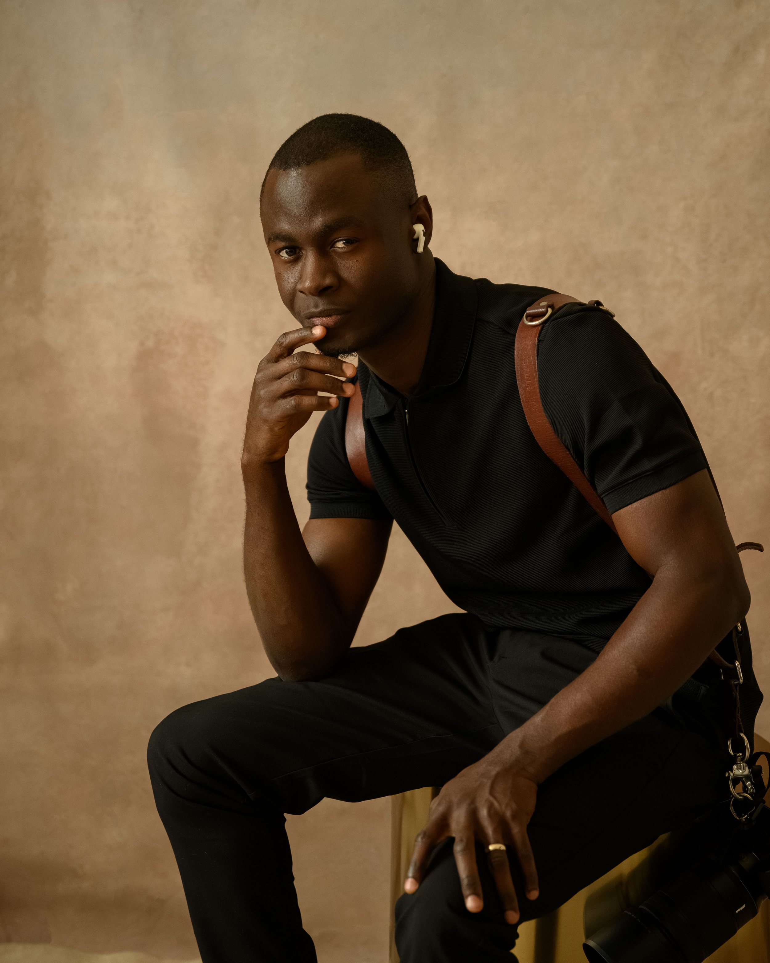 A man sitting on a stool wearing a black polo shirt and black pants, with a brown backpack, white wireless earphones, and a ring on his right hand, posing with his left hand resting on his knee and his right hand near his chin, against a beige background.