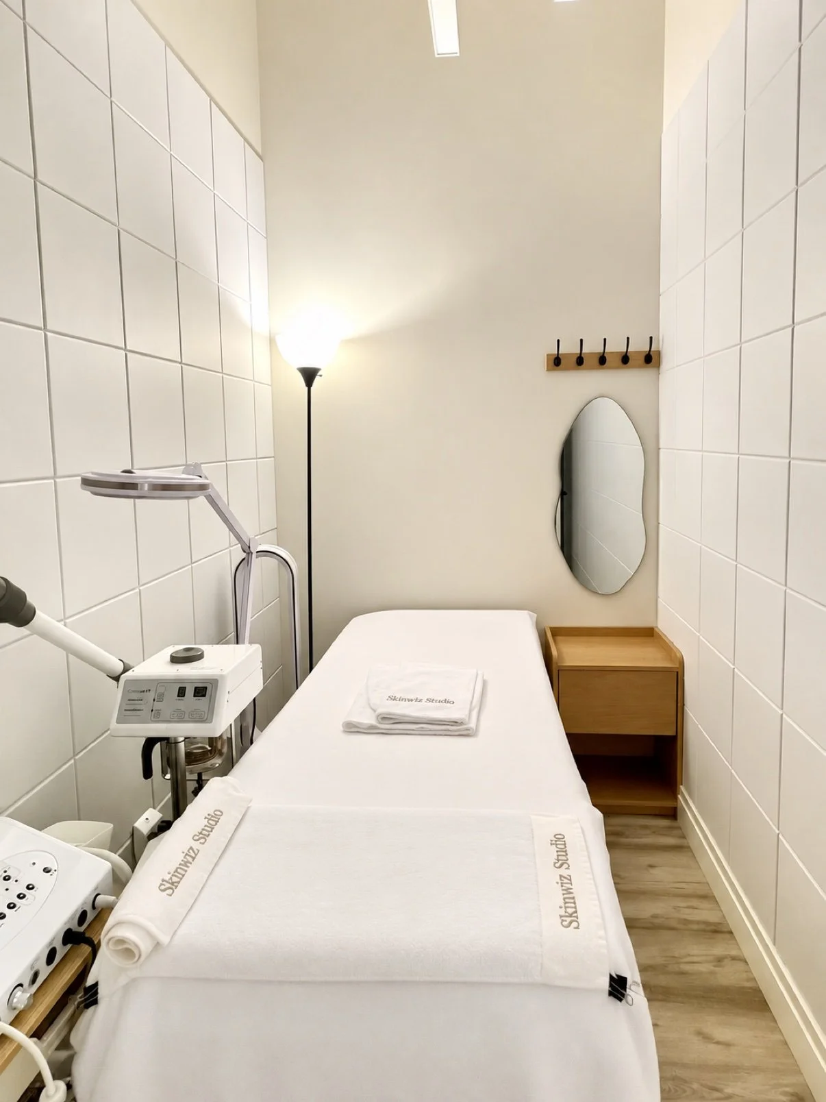 Skinwiz Studio treatment room with white facial bed, branded towels, warm floor lamp, wooden nightstand and mirror in Midtown Manhattan