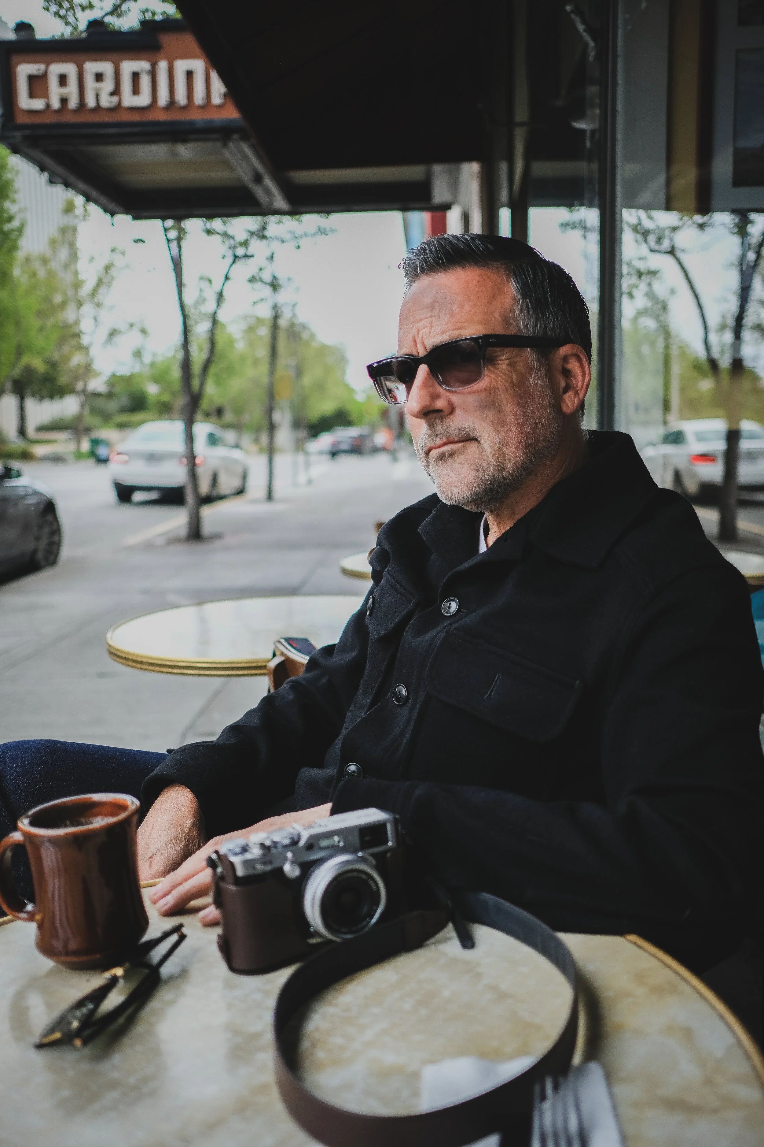 A man wearing sunglasses and a black jacket sitting outside at a cafe table with a cup of coffee, a camera, and eyeglasses placed on the table.