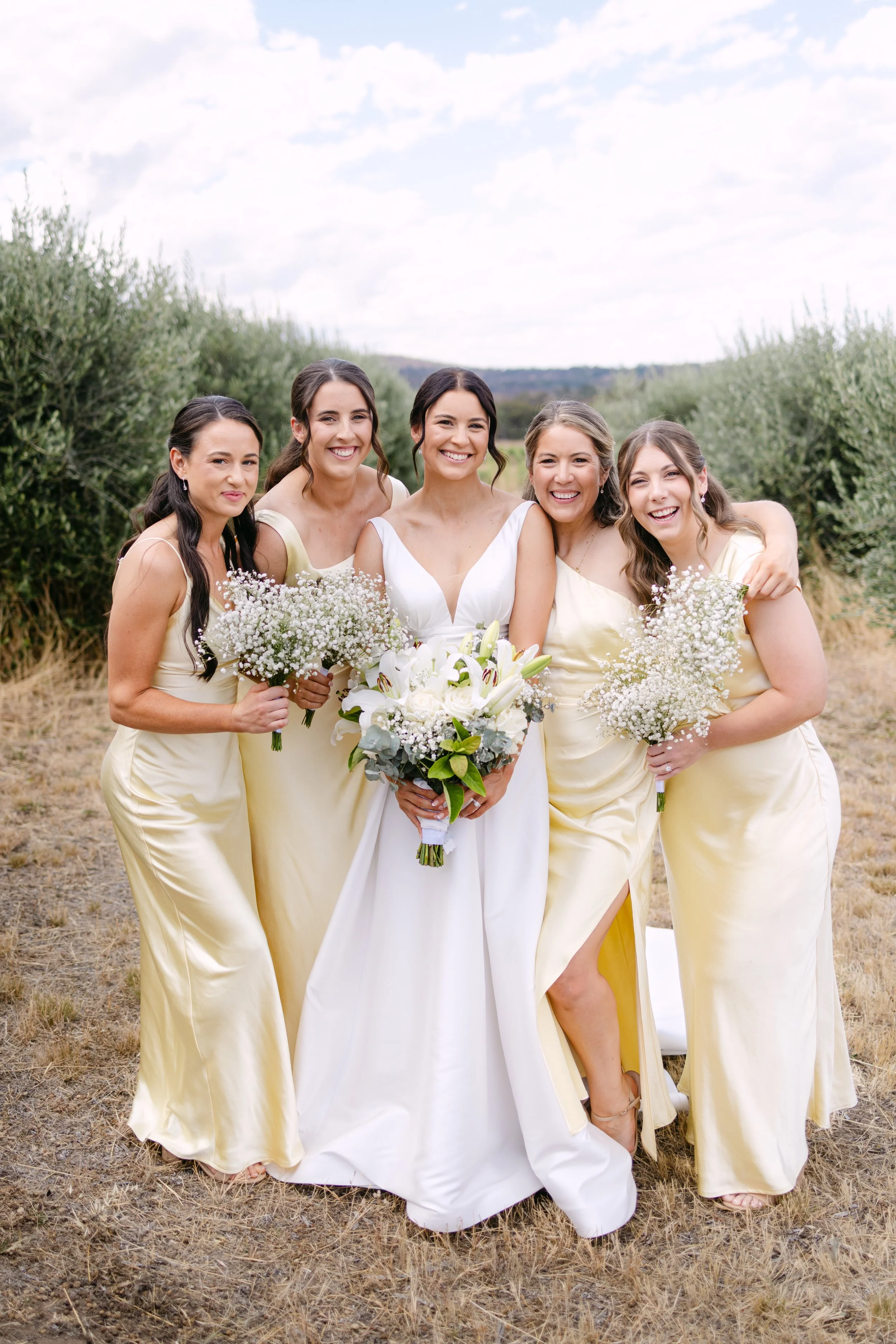 A bride in a white dress holding a bouquet, with five bridesmaids in matching yellow dresses holding bouquets, standing outdoors in a grassy field with trees and hills in the background.