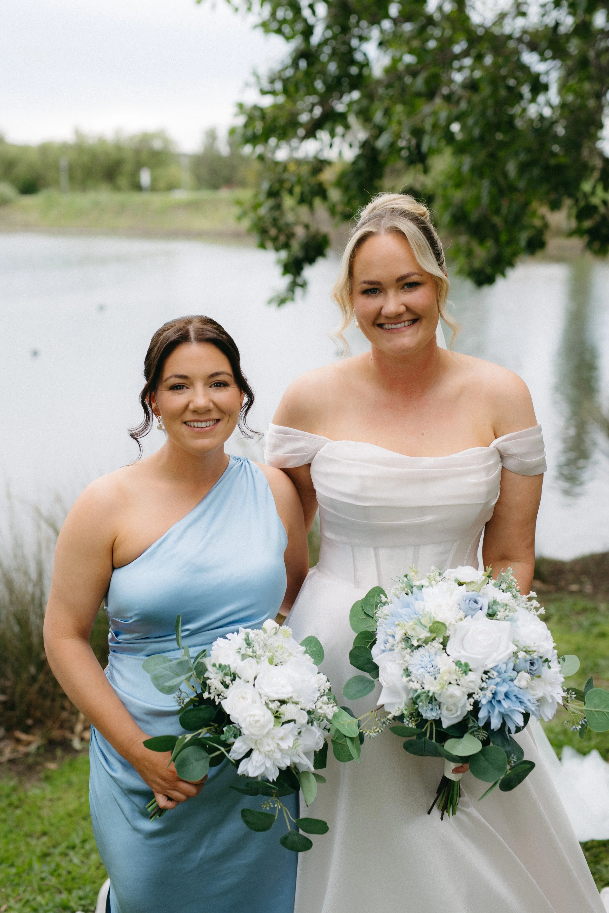 Two women, one in a white wedding dress and the other in a light blue dress, standing outdoors near a lake with trees in the background, holding bouquets of white and blue flowers, smiling at the camera.