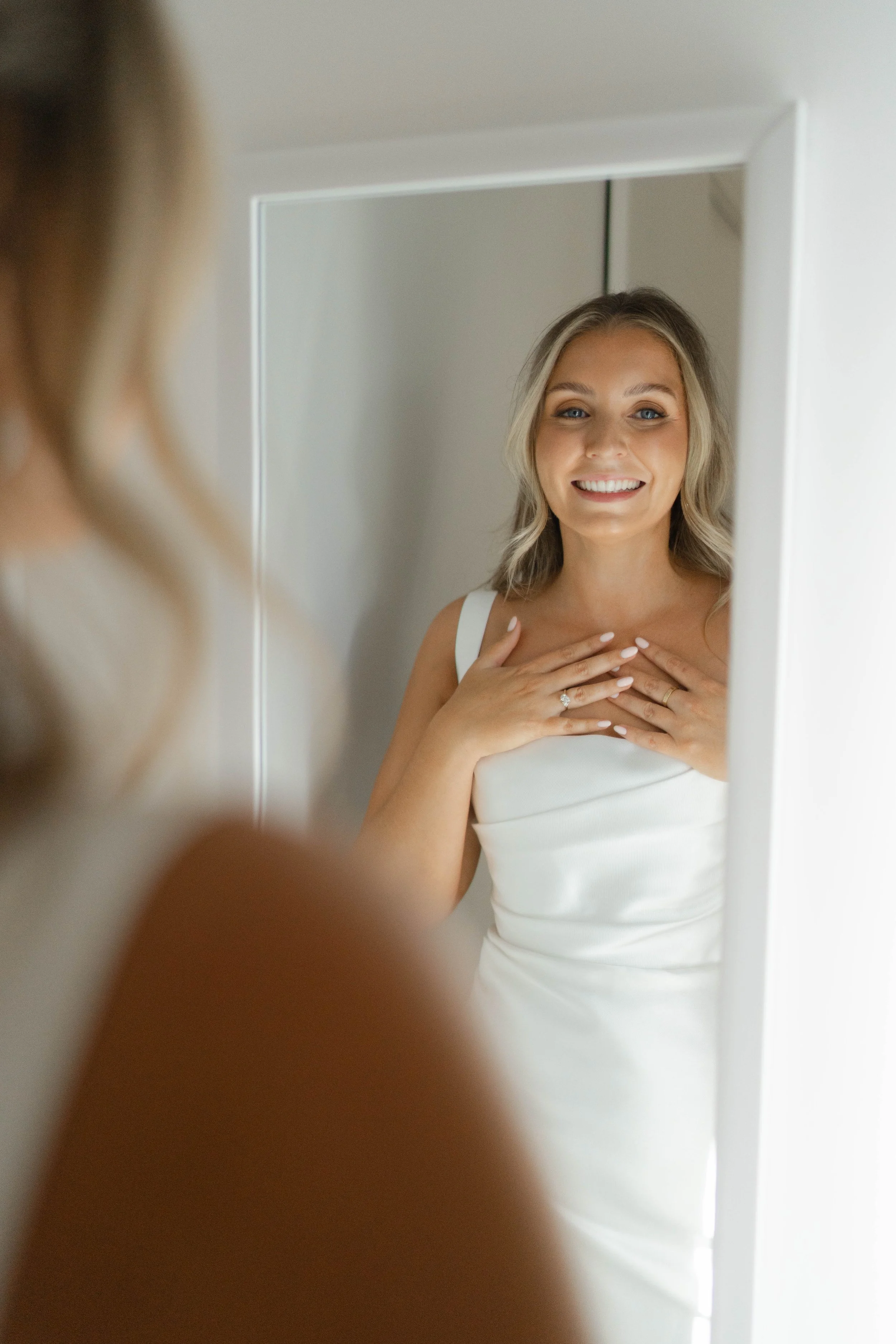 Woman in a white dress looking at herself in a mirror with a smile.