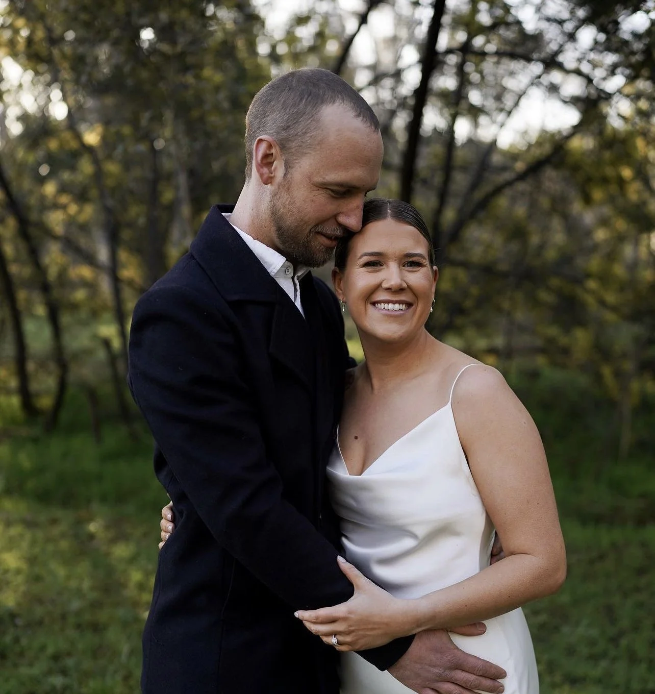 A happy couple embracing outdoors in a park during daytime, smiling at the camera. The woman is wearing a white dress and the man is wearing a dark suit.