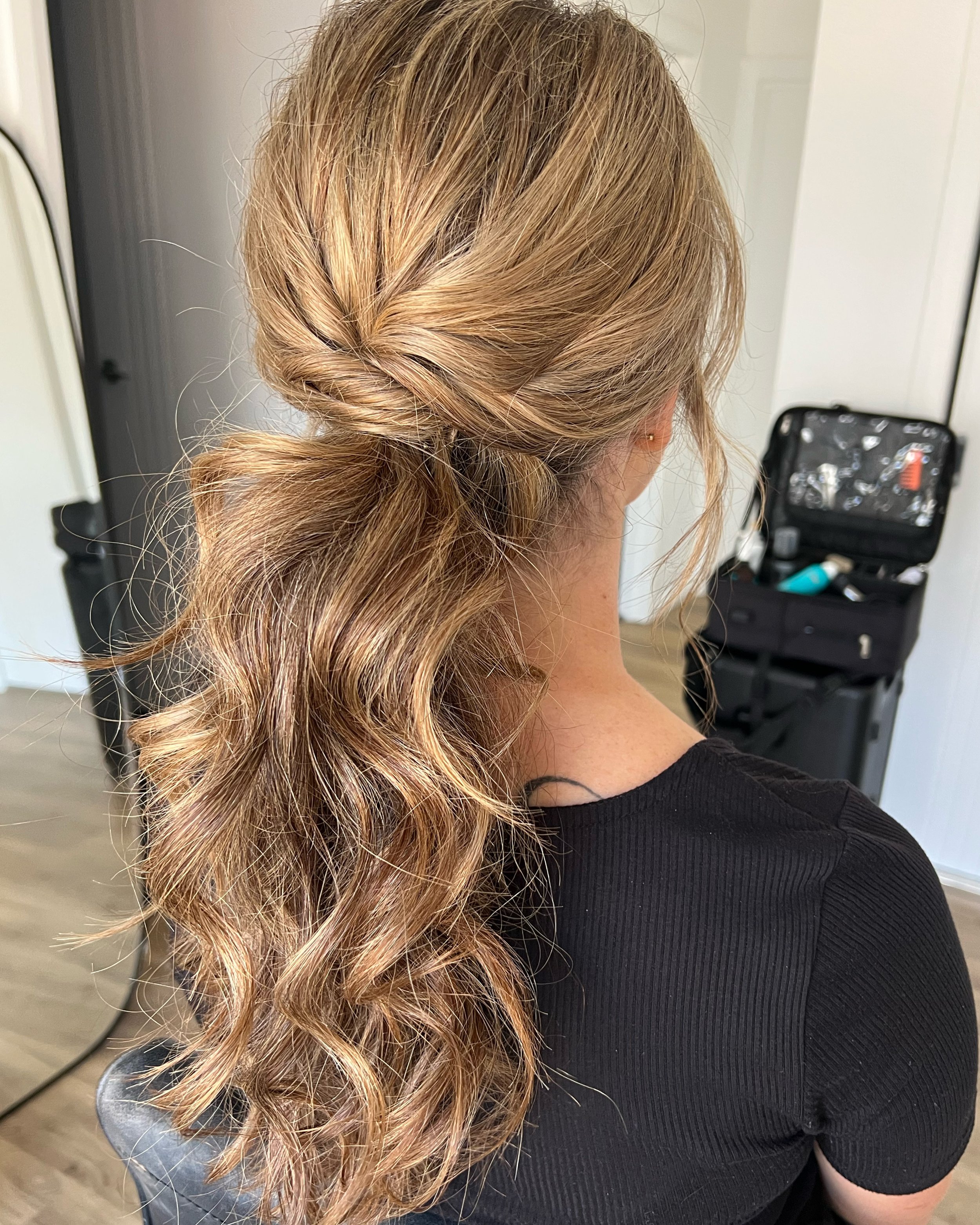 Back view of a woman with long, wavy, light brown hair styled in a loose braid on top, with the rest flowing in waves down her back, wearing a black top, in a room with a hairstyling kit in the background.