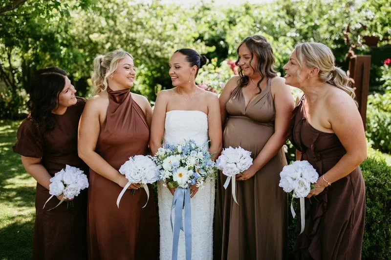 A bride and four bridesmaids standing outdoors in a garden, smiling and holding bouquets, during a wedding celebration.