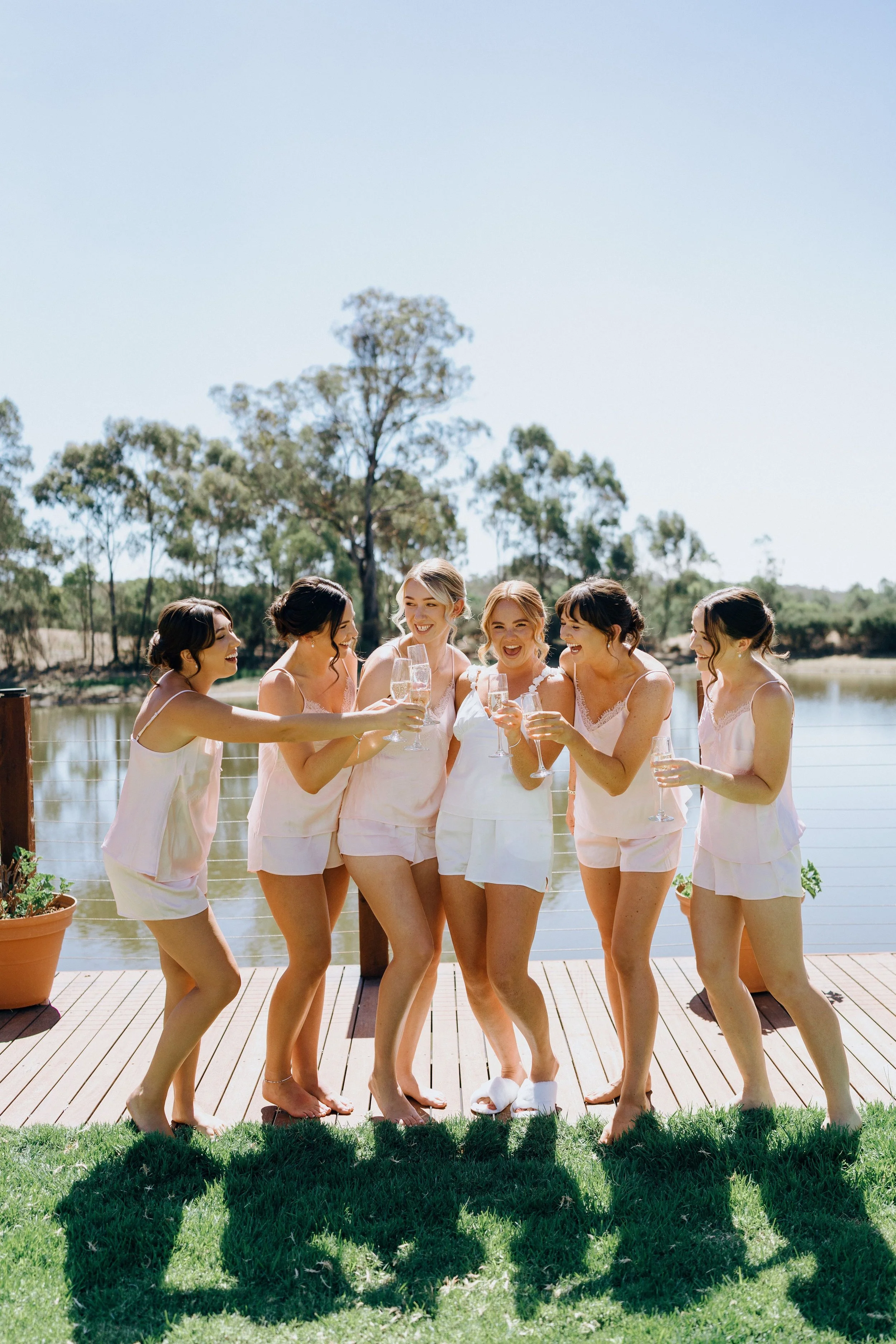 Six women in matching sleepwear celebrating with drinks on a deck by a lake.