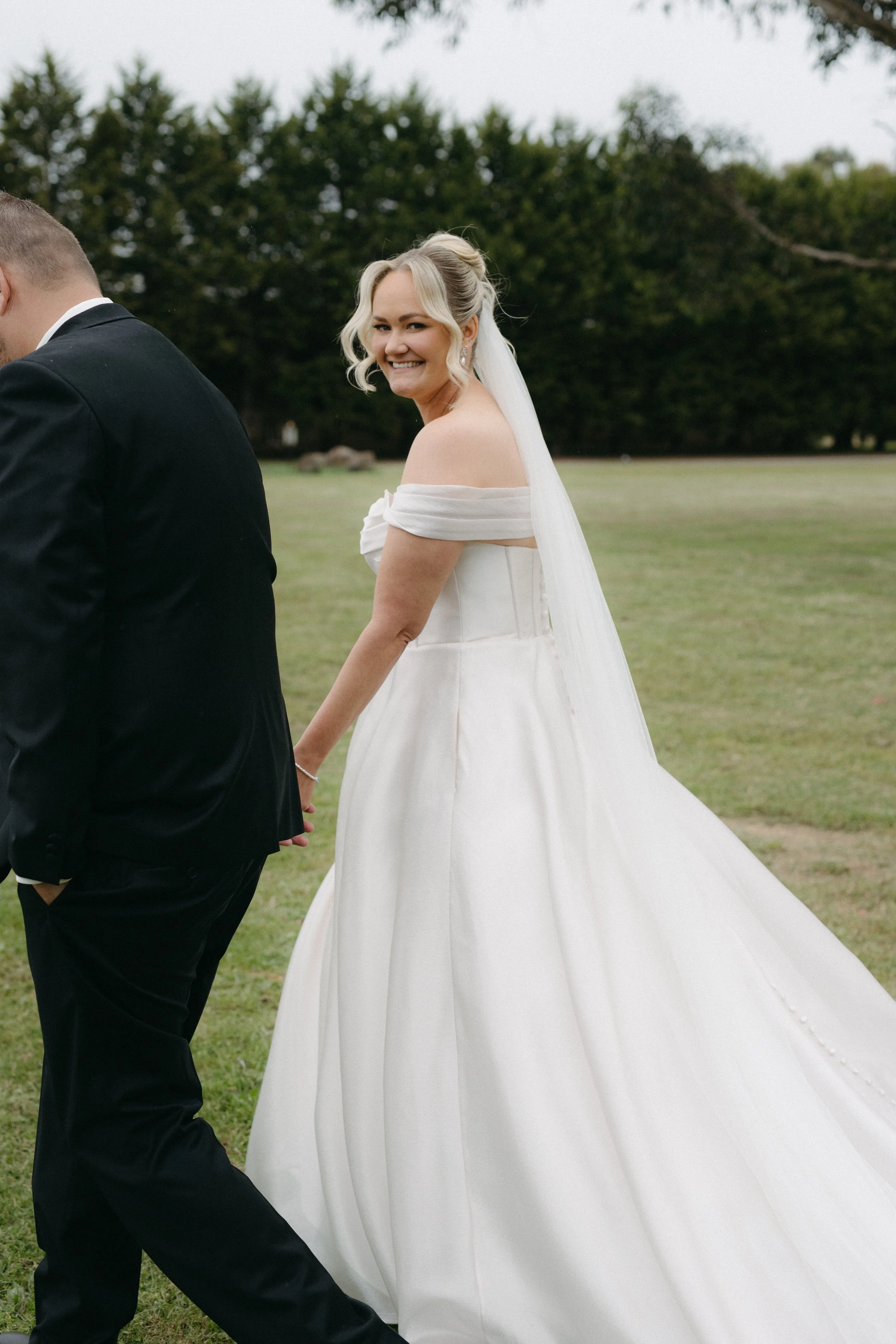 A smiling bride in a white off-the-shoulder wedding dress holding hands with a groom in a black suit outside on a grassy field.