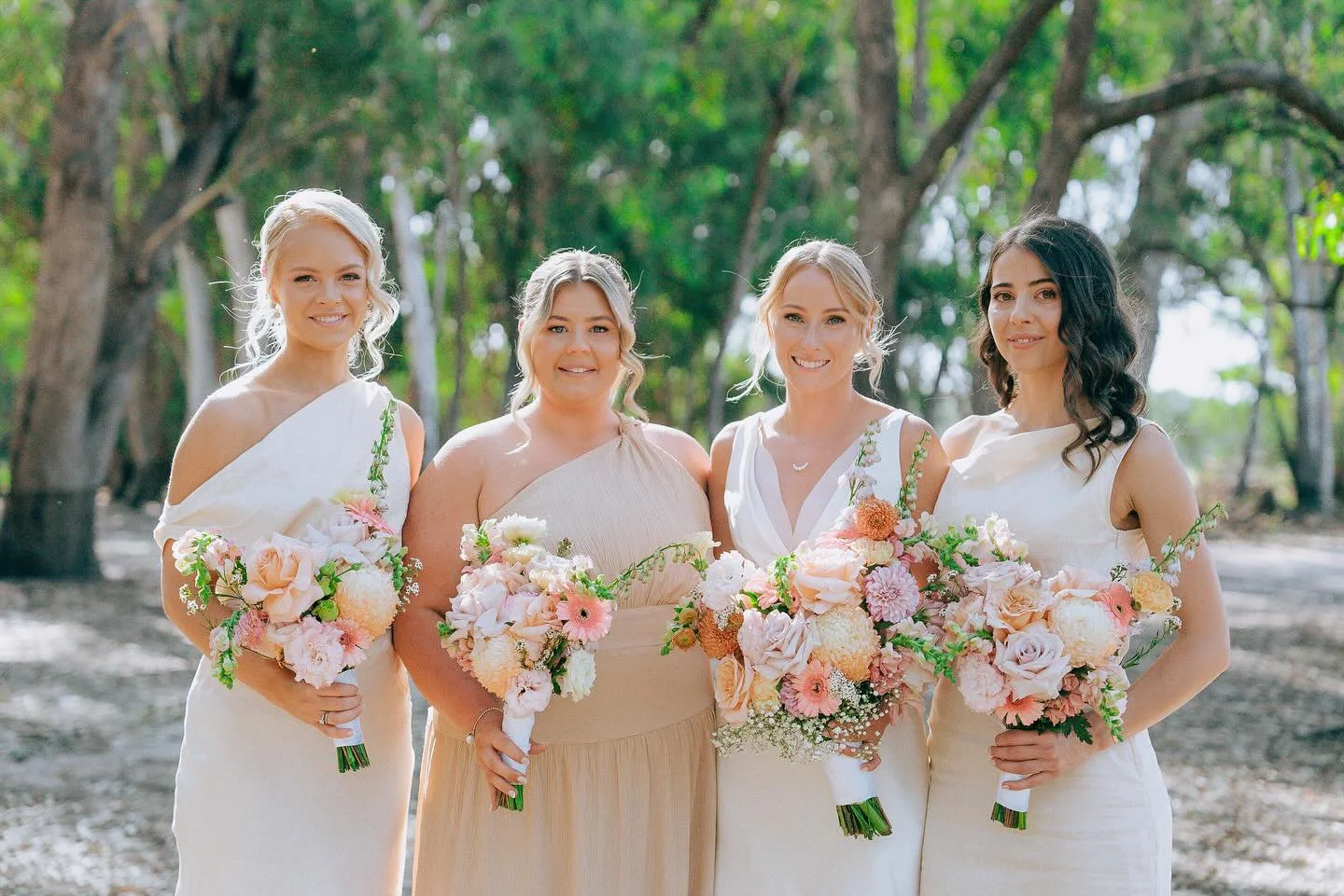 Four women in light-colored dresses holding bouquets of pink and white flowers, standing outdoors in a wooded area.