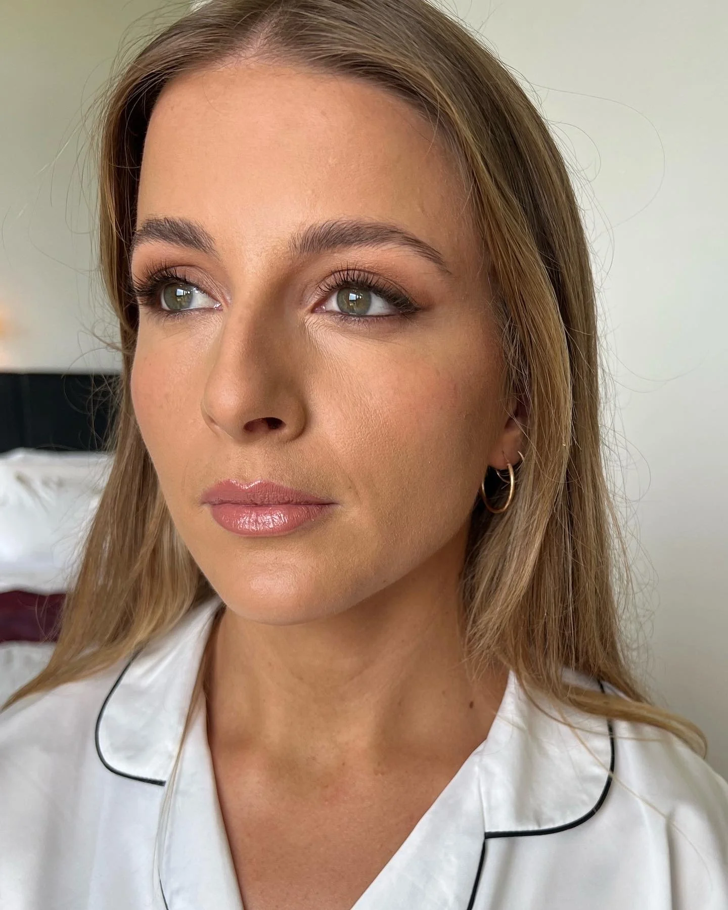 Close-up portrait of a woman with light brown hair and blue eyes, wearing a white blouse with black piping and small hoop earrings, looking slightly to the side.