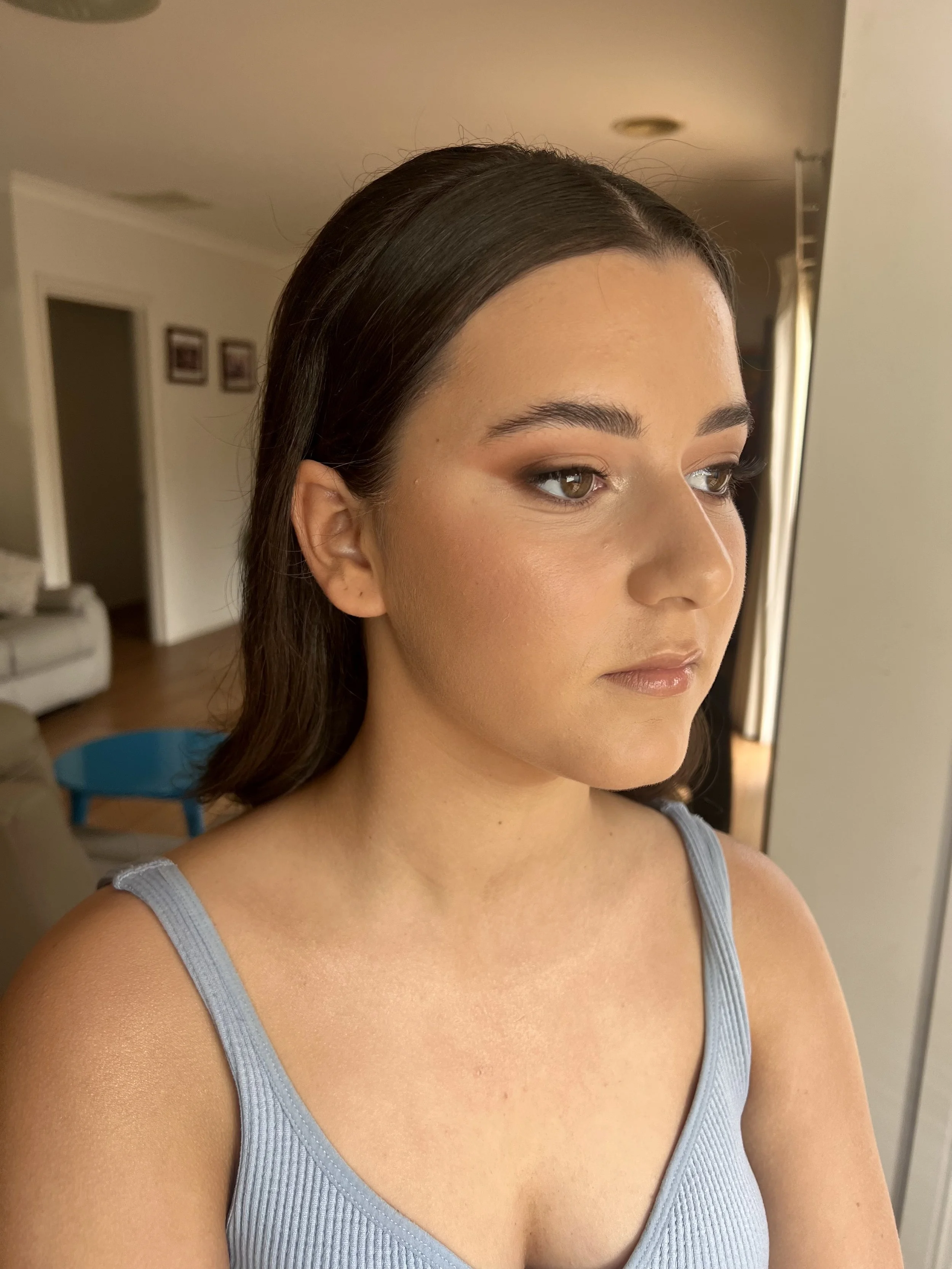 A young woman with brown hair and neutral makeup, wearing a light blue tank top, looking slightly to the right in a warmly lit room.